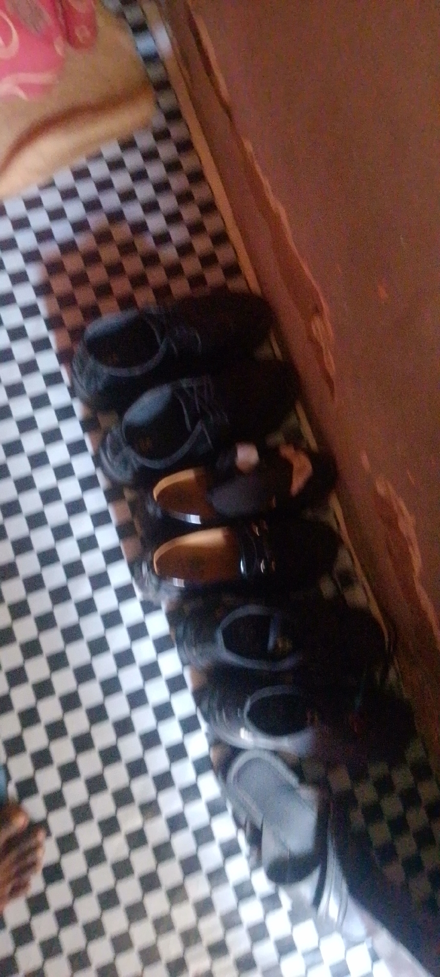 An overhead shot shows a row of shoes placed on a black and white checkered floor. The shoes are all dark in color, with several pairs of black dress shoes, some with a visible tan accent. There are also a few pairs of dark slip-on shoes, some of which appear to be made of a shiny material. The shoes are arranged in a line, with the toes of some pointing towards the viewer and the heels of others. The background is blurry, but appears to be a wall with a reddish-brown hue and a hint of fabric that is pink and tan. The scene is indoors, likely a domestic setting, possibly a hallway or entryway. The image quality is low, with a grainy texture and poor lighting, making it difficult to discern finer details. The location is specified as Jalingo, Nigeria.