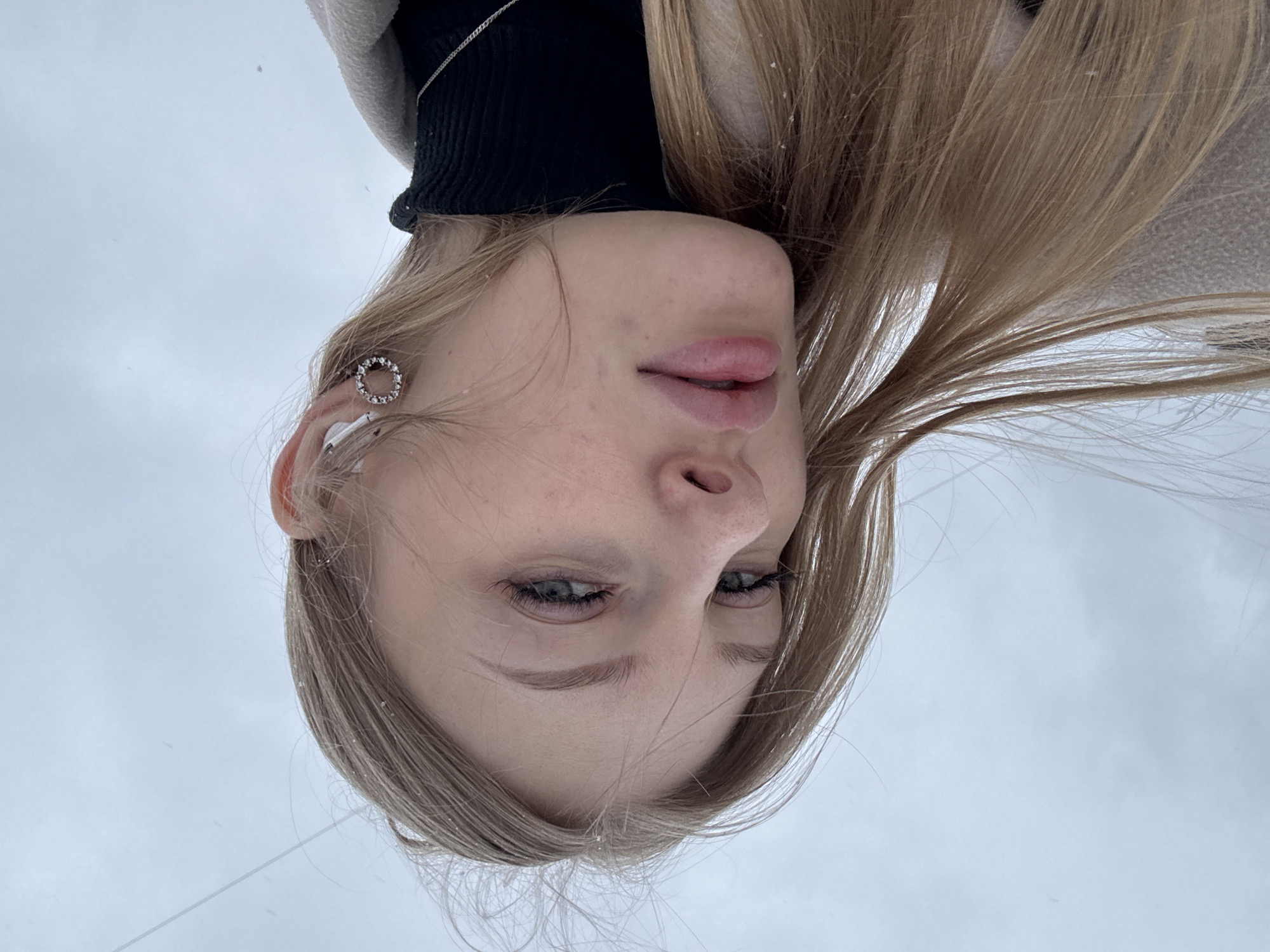 The image is a close-up, tilted portrait of a blonde woman with fair skin and grey eyes. She is wearing a black turtleneck sweater and light-colored outer garment. Her hair is long and straight. She has a silver hoop earring in her left ear. The background is a blurred, pale sky, suggesting outdoor lighting. Fine details suggest snowflakes on the subject's hair and clothing. The location, Perm, Russia, is not definitively discernible from the image content.