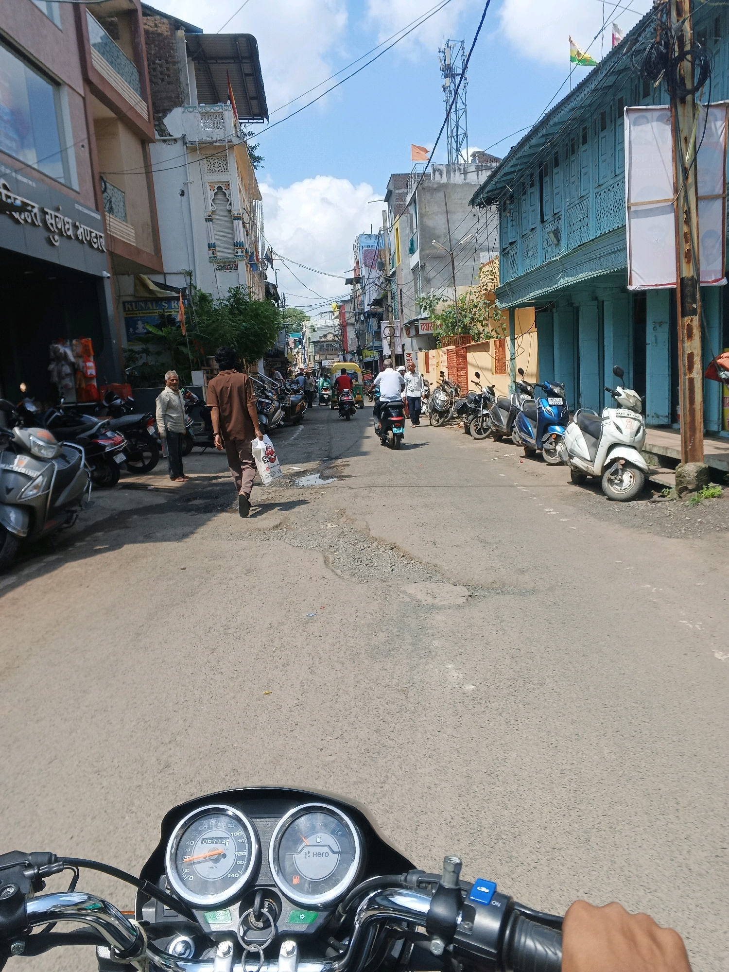 The image depicts a street scene in Dewas, India, on a bright, sunny day with scattered clouds. The perspective is from a motorcycle, showing its handlebars and dashboard in the foreground. The street is paved with asphalt and appears somewhat worn.
On the left side of the street, there are multi-story buildings with shops at ground level. One building prominently displays text in Hindi, including "सन्तोश संग्रह भण्डार". Several parked motorcycles and scooters are visible along the sidewalk. A man in a light-colored shirt and dark pants stands near these vehicles, facing away from the camera.
Further down the street, on the right, is a building with traditional blue shutters and ornate architectural details. Flags are visible on top of some buildings, including what appears to be the Indian flag and other regional flags. A traffic light is visible in the distance.
Several people and vehicles are present on the street. A man in a brown shirt and pants is walking away from the camera towards the center of the street, carrying a white plastic bag. Other individuals are seen on motorcycles and scooters, some moving and some parked. A yellow auto-rickshaw is also visible further down the road. The overall environment suggests a busy urban street, likely in a commercial area.