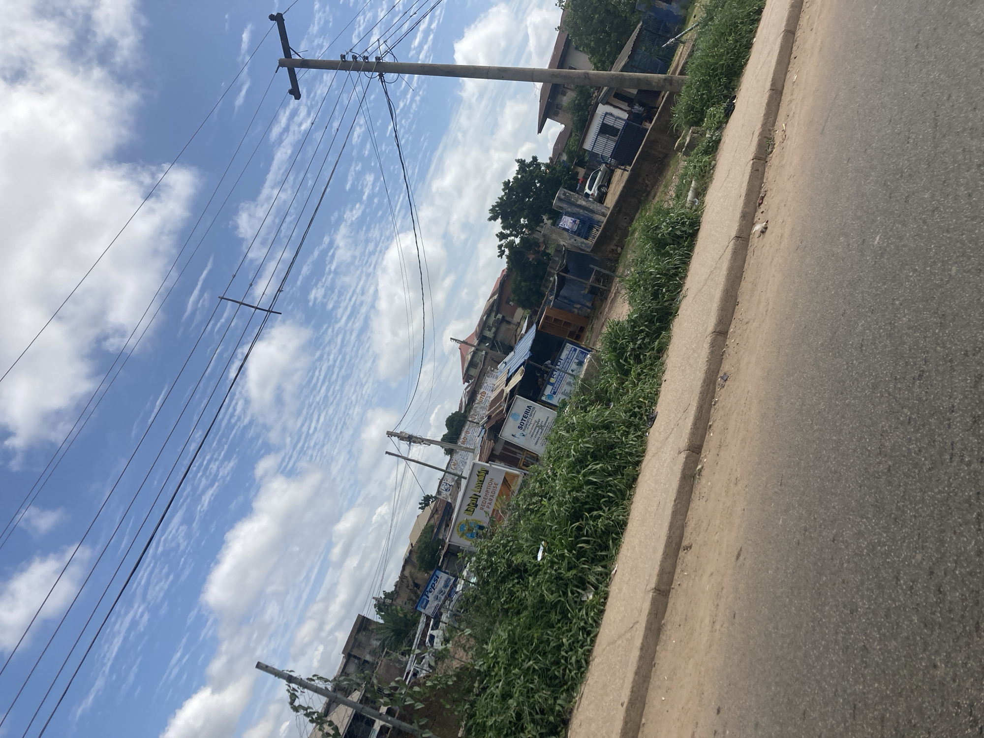 The image depicts a street scene in Jos, Nigeria, under a partly cloudy sky. A paved road occupies the lower portion of the frame, with a concrete curb and a strip of overgrown green vegetation bordering it. Beyond the vegetation, a row of small buildings and structures is visible, some with tin roofs. A prominent utility pole with multiple wires extends vertically along the right side of the image. Several signs are affixed to the buildings, including one advertising "Pepsi." In the middle ground, there is a silver vehicle parked. The background features more buildings and trees, indicative of an urban or semi-urban environment. The overall impression is that of a typical commercial or residential street in Jos.
