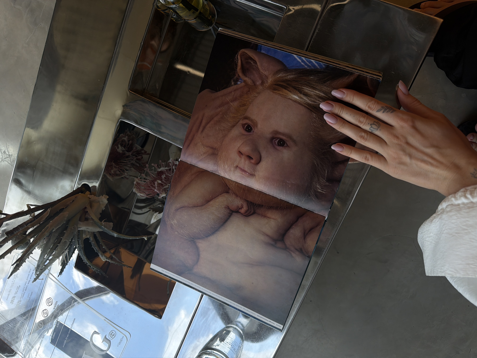 In North Kuta, Indonesia, a person's hand with a lotus tattoo on the ring finger is placed over an open book on a polished metallic surface. The book displays a striking image of what appears to be a piglet's face, its eyes looking directly at the viewer, with delicate, human-like skin. The background of the book's image is a blurred blue and white striped pattern. The metallic surface reflects the environment, including a clear bottle with a dark liquid, possibly oil, and an abstract dried plant arrangement. The overall scene is indoors, with a cool, modern aesthetic.