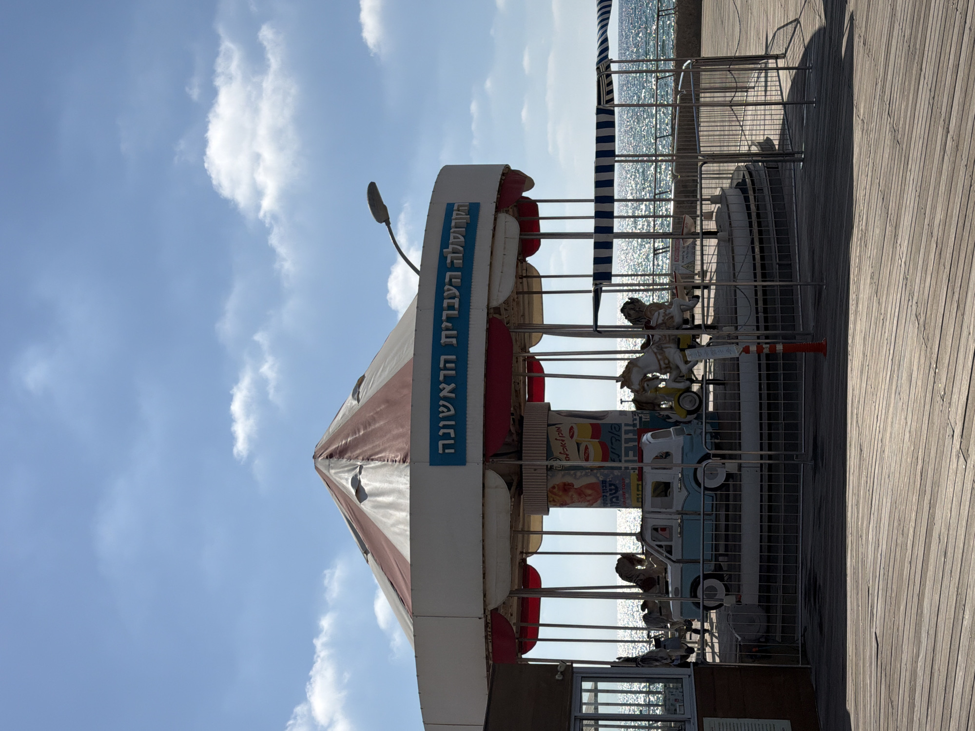 A carousel with a weathered, conical roof of white and maroon stripes stands on a wooden boardwalk beside the shimmering sea under a bright blue sky with scattered clouds. The carousel features several decorative elements, including a light blue and white car, and horses. A blue sign with Hebrew lettering is affixed to the side of the carousel's upper structure. In the background, the sea extends to the horizon, reflecting sunlight. The scene suggests a public amusement area, possibly a boardwalk attraction in Tel Aviv, Israel, given the Hebrew signage.