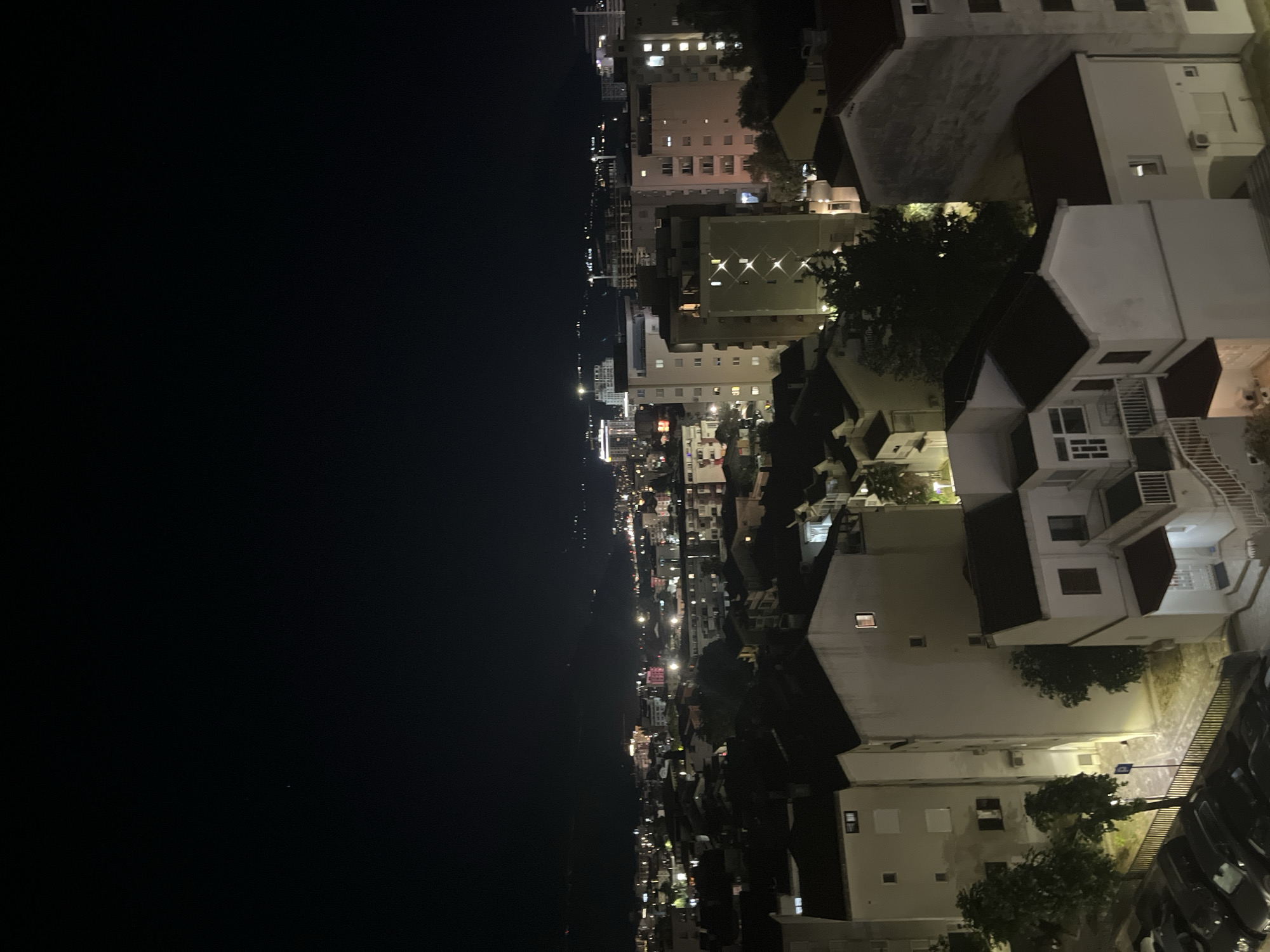 This is a nighttime aerial view of a city, believed to be Budva, Montenegro, showcasing a densely populated urban landscape with a mix of residential buildings and some taller structures in the distance. The sky is dark, with a few faint stars visible. The city lights are illuminated, casting a warm glow across the scene.
In the foreground, several multi-story residential buildings are prominent. These buildings are characterized by their light-colored facades, dark-tiled roofs, and numerous illuminated windows. Some of these buildings appear to be apartment blocks with balconies. In the lower left corner, a parking lot filled with various cars is visible. Adjacent to the buildings, there are some trees, their foliage dark against the night sky, and pathways are illuminated by streetlights.
Further into the background, the cityscape extends, with more buildings of varying heights. Some taller structures, possibly hotels or commercial buildings, are discernible, their windows also lit. The distance is filled with a tapestry of lights, indicating the ongoing activity within the city. The overall impression is one of a lively urban center at night, with a sense of depth and scale created by the receding buildings and the vastness of the dark sky. The scene is devoid of any visible people or specific actions.