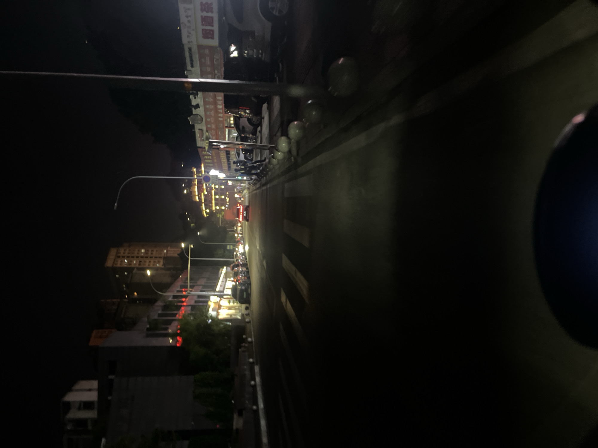 This image depicts a nighttime urban street scene in Jiulongpo District, China. The perspective is low, likely from a vehicle, looking forward down a multi-lane asphalt road. White pedestrian crosswalk markings are visible on the road surface in the foreground.
On the right side of the street, a row of commercial buildings features illuminated storefronts and signage in Chinese characters, prominently displaying "烟酒茶" (Tobacco, Alcohol, Tea) on one awning. A series of white spherical bollards delineate the sidewalk from parked vehicles, which include a white SUV, a dark SUV, and other sedans further down. A tall, thin utility pole stands on the right.
On the left side of the street, several multi-story buildings are largely dark, though some windows and red lights are visible. Further in the distance on the left, a distinct, ornate building complex is brightly illuminated with yellow lights. Multiple streetlights line both sides of the road, casting a mix of white and yellow light across the scene, creating significant shadows. Red taillights from moving vehicles are visible further down the street, indicating active traffic. A blue circular sign is partially visible on the left sidewalk. Indistinct human figures can be faintly observed on the right-hand sidewalk.