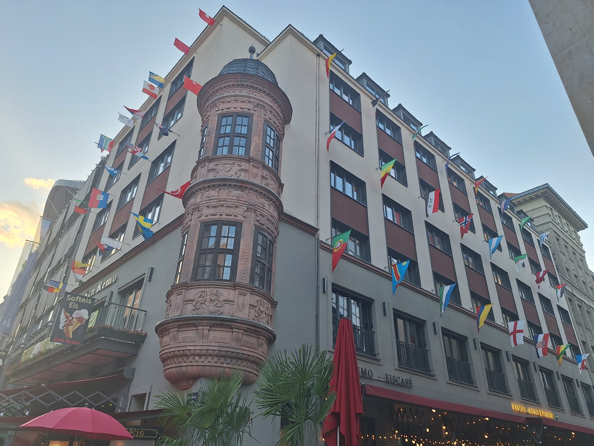 A building in Leipzig, Germany is decorated with numerous small national flags displayed on poles along its facade. The building has a light-colored exterior with dark-trimmed windows and several balconies. A prominent architectural feature is a circular, reddish-brown turret with intricate carvings and a dark, conical roof. A sign on the building reads "San Remo Softeis Eis". Red umbrellas are visible in the foreground, along with palm trees and outdoor seating. The sky above is clear and pale blue.