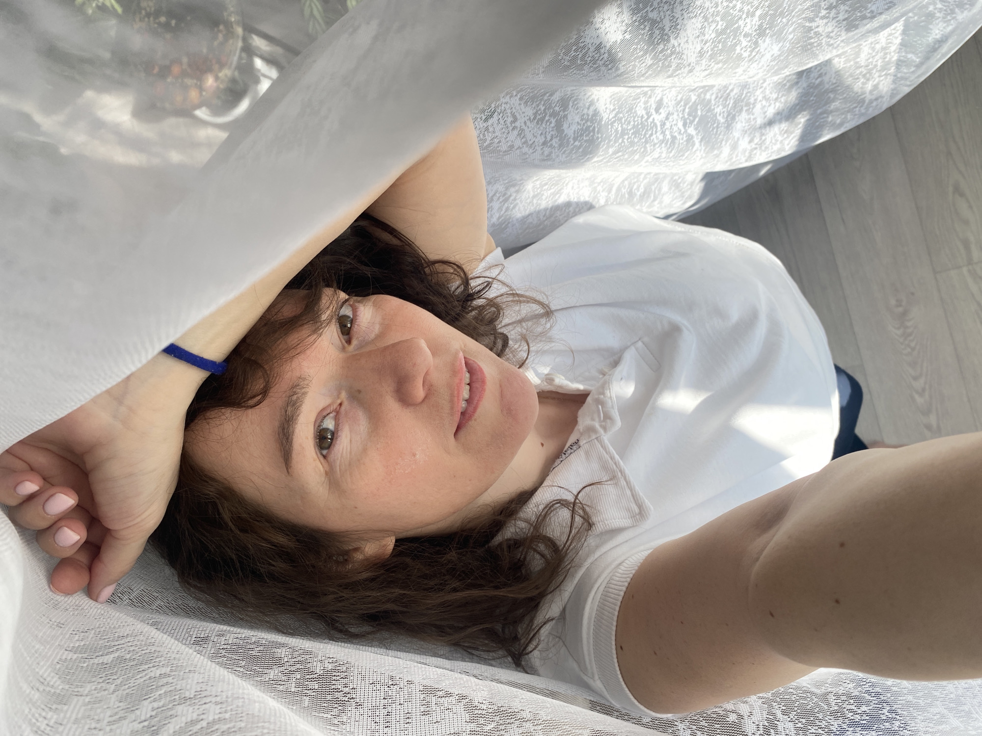 A middle-aged Caucasian woman with brown curly hair and fair skin is lying down, partially obscured by a white lace curtain. She is wearing a white collared shirt and a blue bracelet on her left wrist. Her head is tilted, and she is looking up at the camera with a slight smile. The background includes a light-colored wooden floor and a glimpse of what appears to be a plant in a glass container. Based on the prompt mentioning the city of Ольховка, Russia, the photograph was likely taken indoors in a residential setting in that location.
