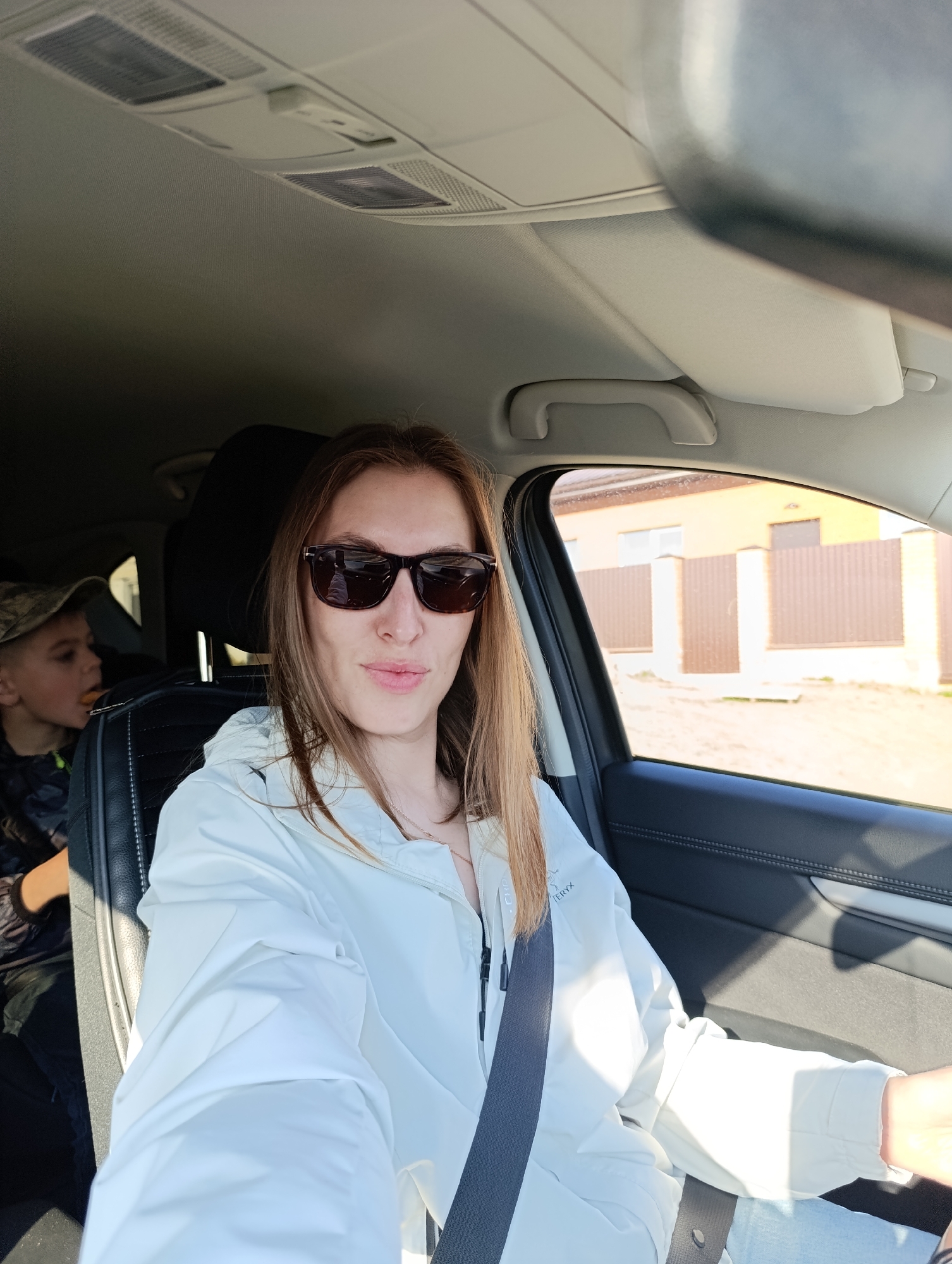 A woman wearing sunglasses and a white jacket is sitting in the driver's seat of a car, with a seatbelt fastened. A young boy with a camouflage hat is in the back seat. The view through the car window shows a fence and a building. Based on visual markers, the location is within the city of Chaikovsky, Perm Krai, Russia.