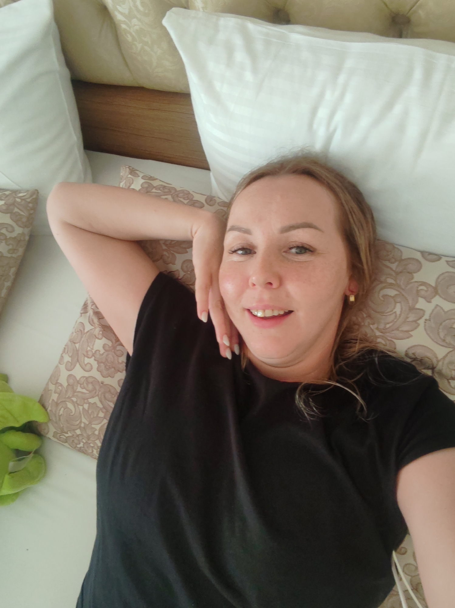 A woman lies on a bed with patterned cushions and white pillows. She wears a black T-shirt and is smiling, with one hand near her face. The location is Alanya, Turkey.