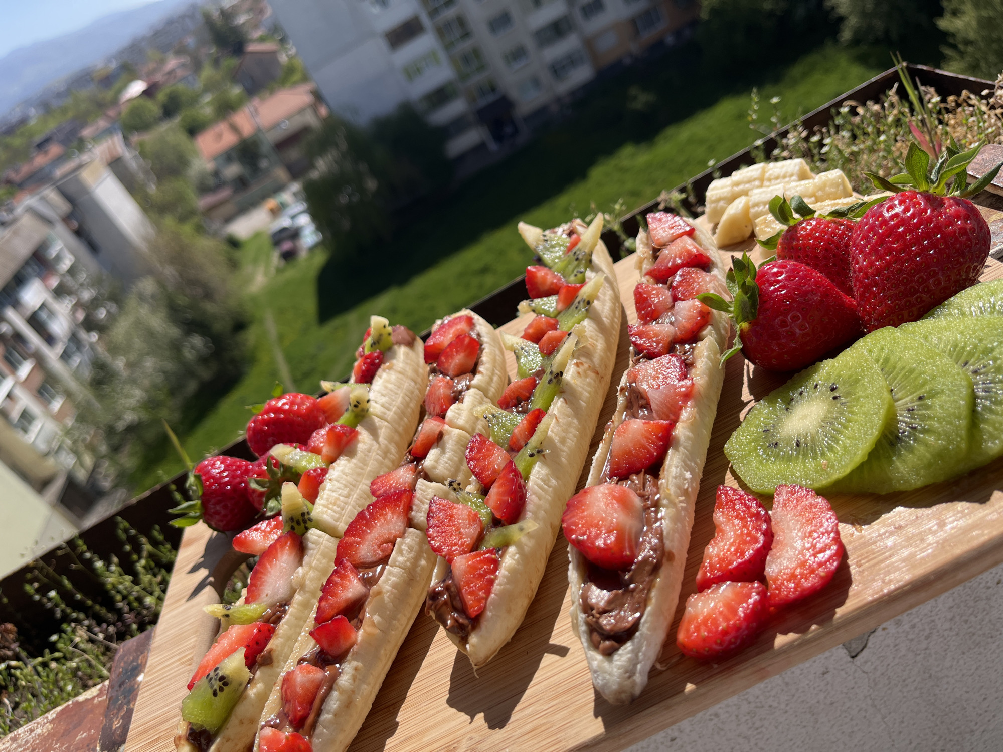 The image displays a close-up, high-angle shot of several fresh fruit snacks arranged on a light-colored wooden cutting board. The main subjects are five peeled bananas, each split lengthwise and filled with a dark, creamy spread, likely chocolate or hazelnut, then generously topped with diced fresh strawberries and small pieces of kiwi. Additional whole strawberries, sliced kiwi, and a pile of sliced banana pieces are also present on the board.
The setting appears to be an outdoor balcony or terrace, with a dark metal railing visible behind the food. Beyond the railing, a vibrant green area with grass and trees can be seen. In the background, there are multi-story residential buildings with balconies, typical of an urban or suburban environment. Further in the distance, a blurred mountainous or hilly landscape is visible under a clear, bright blue sky.
The activity depicted is the presentation or preparation of a healthy and colorful fruit-based snack. The strong, direct sunlight suggests it is daytime, likely morning or early afternoon, with clear weather. The light casts distinct shadows on the wooden board and enhances the vibrant colors of the fruit. There are no people visible, nor any discernible text in the image. The architecture and distant landscape are consistent with the location context of Sofia, Bulgaria.