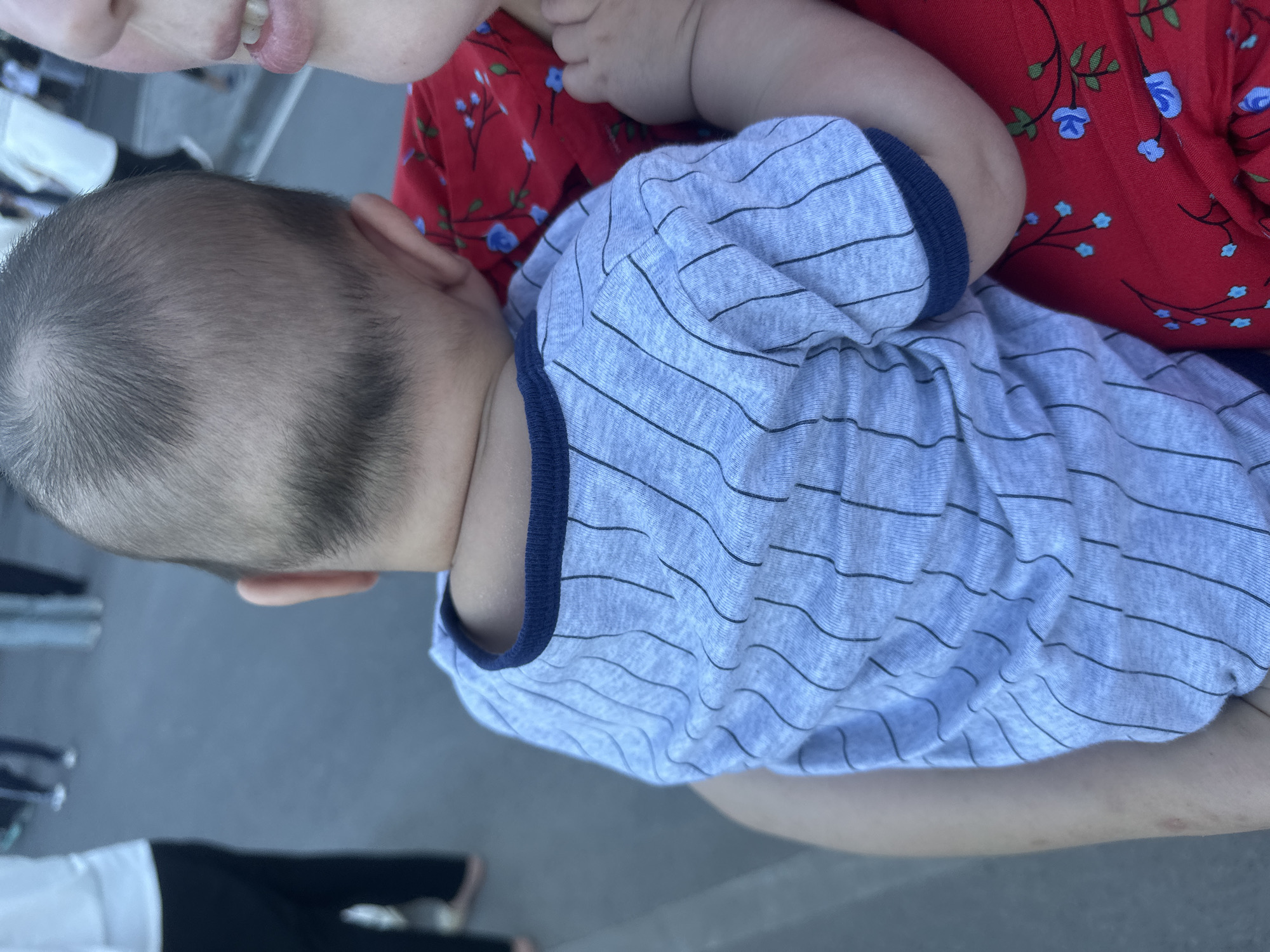 The media file shows a close-up, vertically oriented shot of two people, likely a parent and child, with a blurred background. The focus is on the upper body of a baby wearing a light blue, striped t-shirt with a navy blue trim. The baby's head is turned to the side, revealing their ear and the back of their head, which has short, dark hair. Above the baby, a person's mouth is visible, with lips slightly parted, suggesting an interaction like a kiss or talking. This person is wearing a red top with a floral pattern of blue flowers and green leaves. The setting appears to be outdoors, with a grey, textured surface visible in the background, possibly pavement or a similar ground material. The lighting suggests it is daytime, but no specific weather cues are evident. The overall mood seems intimate and tender, given the proximity of the two individuals. There is no visible text in the frame.