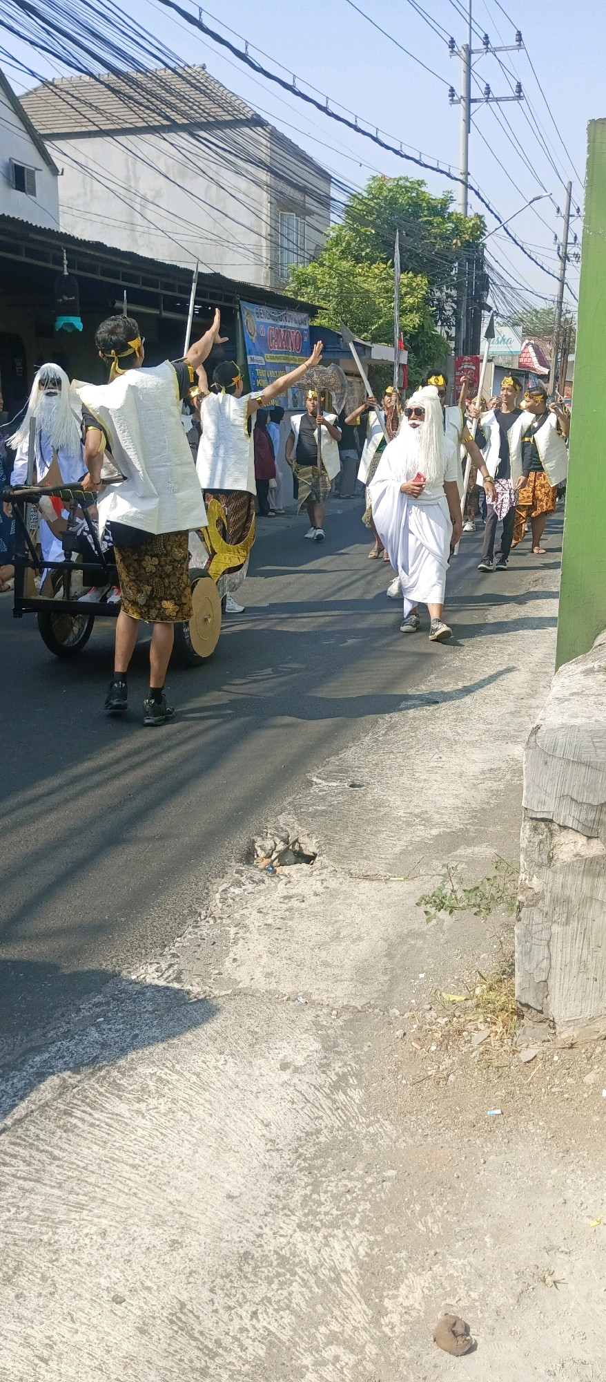 A procession is taking place on a street in Malang, Indonesia, as indicated by the presence of a sign with the word "CAZINO" which is a known establishment in the city, and a prominent utility pole that is characteristic of the infrastructure in Malang. Several individuals are participating in the procession, some dressed in traditional attire with white robes, long white beards, and headbands, while others are wearing white tops with patterned sarongs and golden headbands. Many are carrying long poles or spears, suggesting a ceremonial or performance context. One person in the foreground is standing on a cart, his back to the camera, with arms raised. Another individual, also in white robes and a beard, is walking forward, holding a red object, possibly a phone, in his hand. The environment is urban, with buildings and overhead power lines visible. The road surface is asphalt, with some cracked concrete on the side. The weather appears to be sunny, casting shadows on the ground.