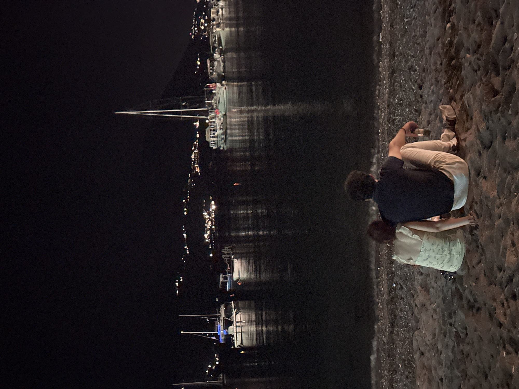 A couple sits on a pebble beach at night, facing a calm bay filled with moored boats. In the background, a hillside town with glowing lights stretches across the horizon, with a prominent mountain peak rising behind it. The water reflects the lights of the town and the boats. The couple is sitting close together, with the woman in a light-colored dress and the man in a dark shirt and light pants. The man is holding a bottle. The overall atmosphere is peaceful and romantic. This scene is likely in the vicinity of Budva, Montenegro, known for its beautiful coastline and vibrant nightlife.
