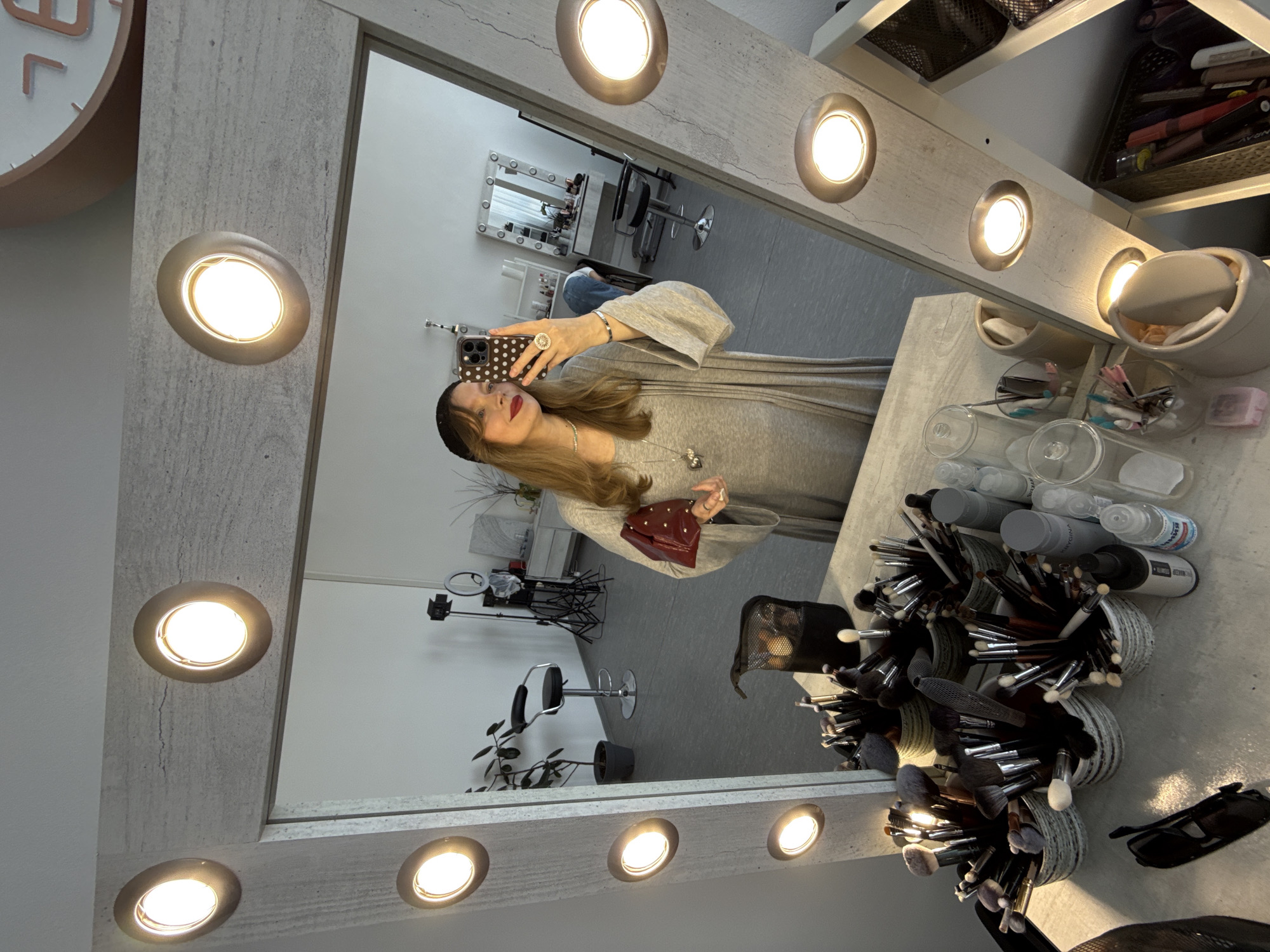 A woman is taking a selfie in a mirror with vanity lights. The reflection shows a makeup station with brushes, cosmetics, and studio equipment in what appears to be a professional makeup setting or salon. The reflection in the mirror shows another mirror in the background. The image does not offer enough information to determine the specific location within Izhevsk, Russia.