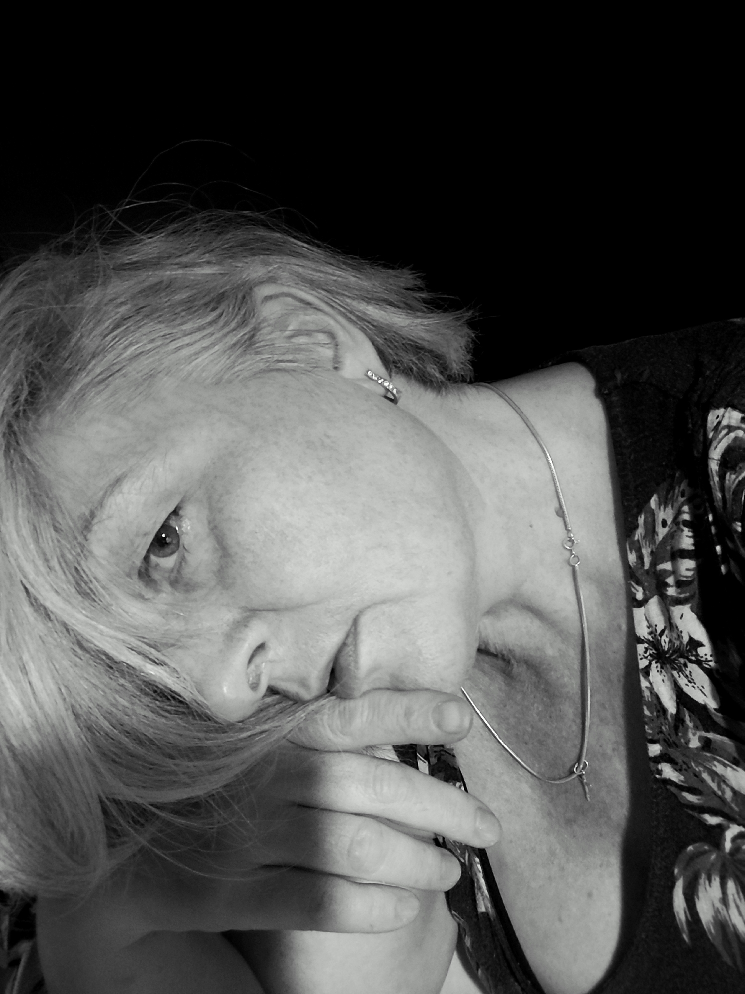 This is a black and white, medium close-up, indoor portrait of a woman. She is leaning forward, with her head tilted and her right hand partially covering her mouth. Her eyes are open, and she appears to be looking directly at the camera. She has short, light-colored hair and is wearing a necklace with a small pendant. Her clothing has a floral pattern. The background is dark and out of focus, suggesting a studio setting or a dimly lit room. The lighting highlights her face and hair, creating a dramatic effect. The overall mood is contemplative or perhaps melancholic. No visible text is present in the image. The time of day, weather, and specific location cues are not discernible from the image itself, though the provided context suggests the location is Krasnodar, Russia.