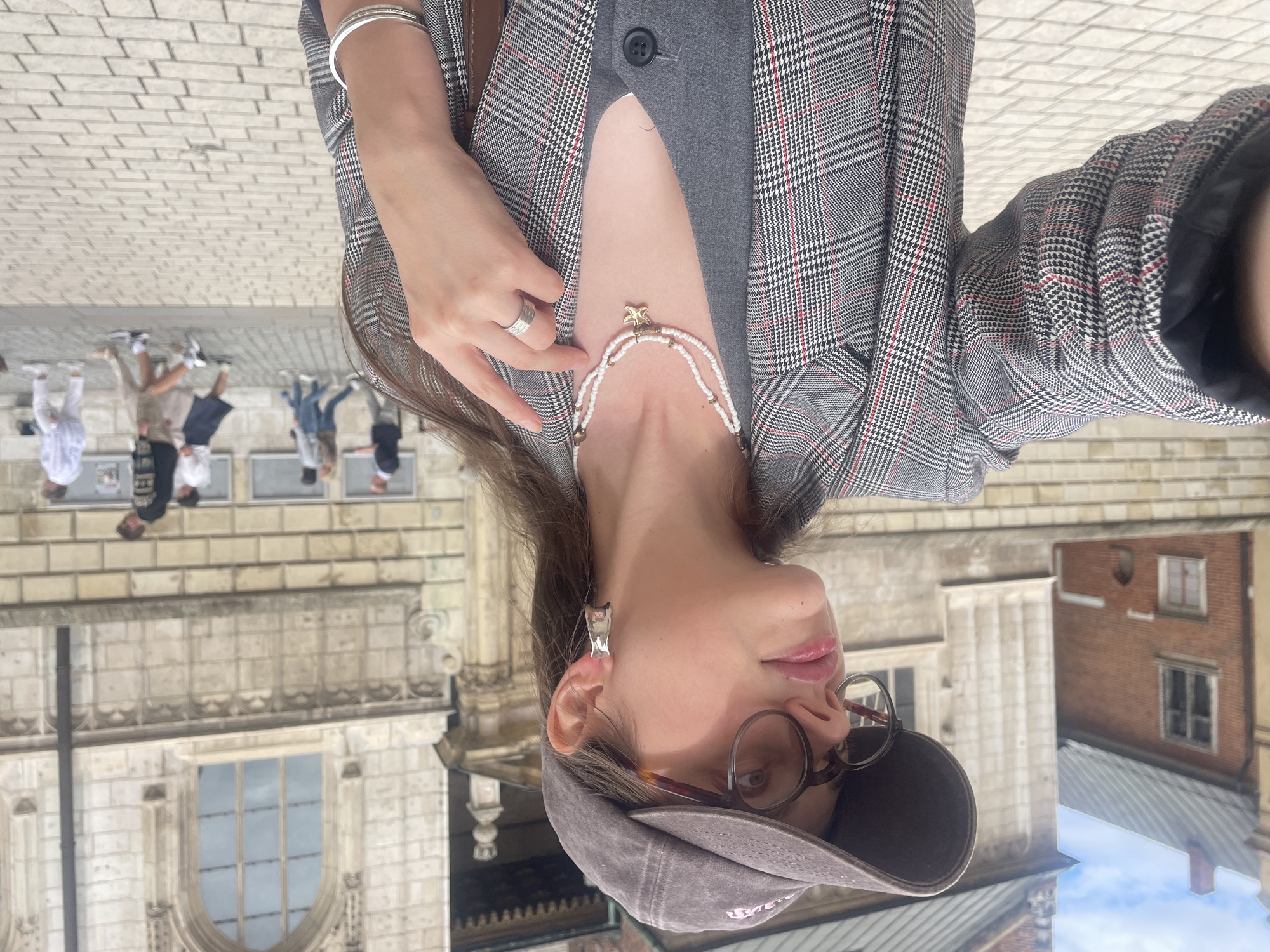 A woman in a plaid blazer and baseball cap takes a selfie in front of a historic building in Krakow, Poland. The woman has on glasses, a necklace with a starfish charm, and a ring. Behind her, several people are walking on a cobblestone street in front of an ornate stone building.