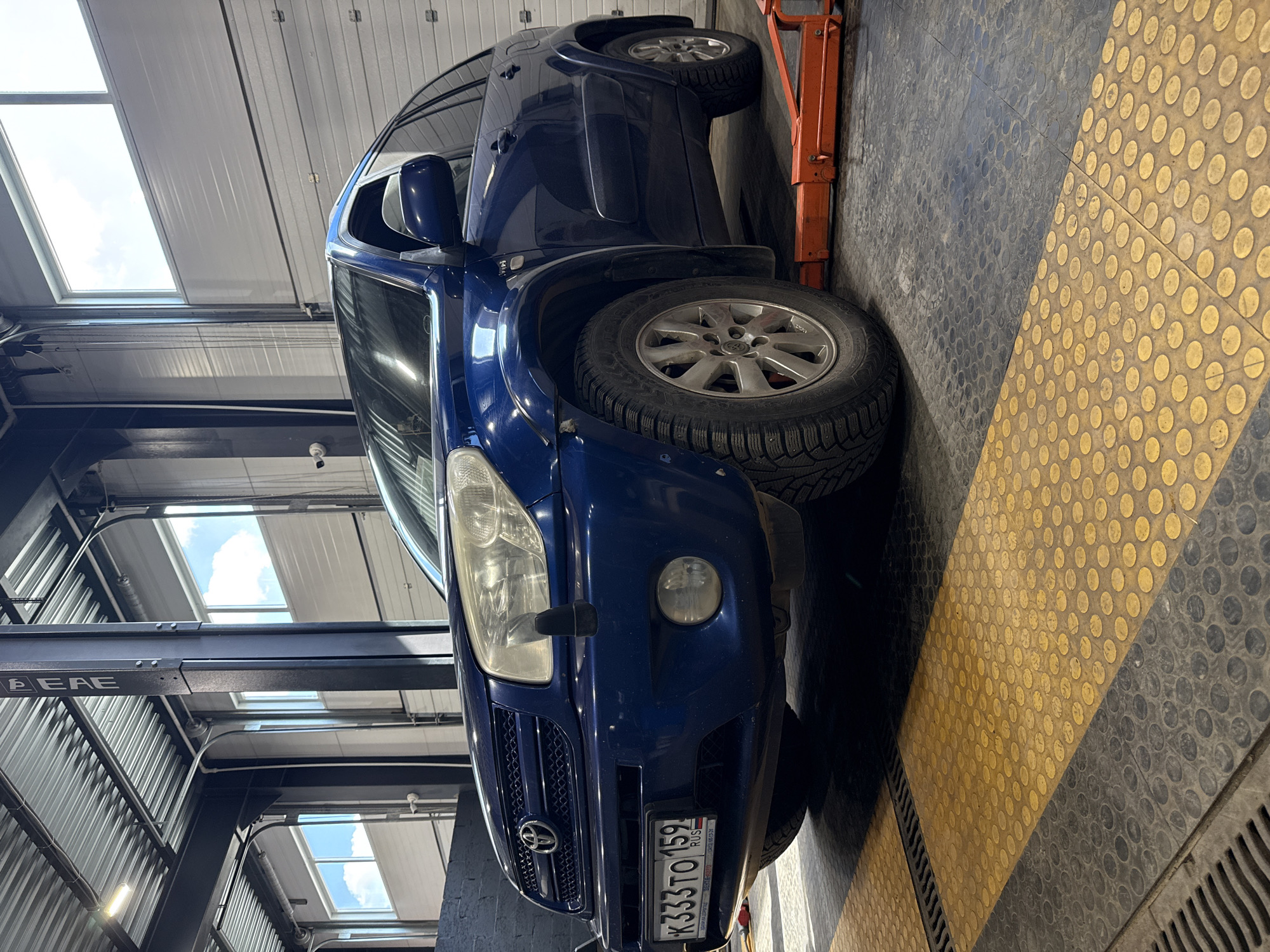 A blue Toyota RAV4 is parked inside a garage-like structure in what appears to be the city of Chaikovsky, Russia, based on the license plate "K333TO159 RUS". The vehicle is positioned on an orange lift, raised slightly off the ground. The floor is covered in a pattern of yellow and gray dotted tiles. The garage structure has a corrugated metal roof with skylights that allow natural light to enter. A surveillance camera is visible on the interior structural beams.