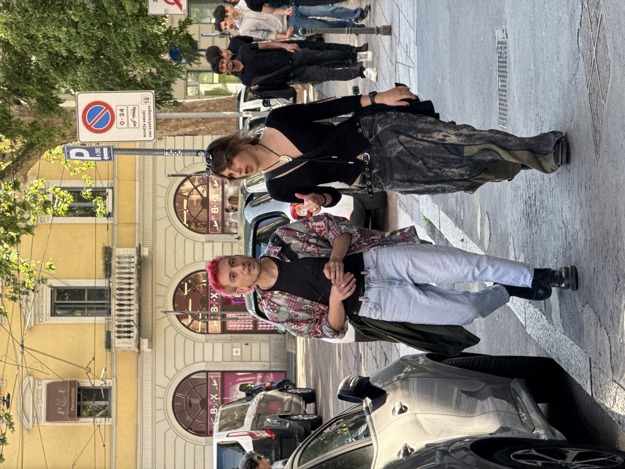 The image, rotated 90 degrees clockwise, captures a vibrant street scene in Milan, Italy, under daylight conditions, indicated by clear shadows. In the foreground, two individuals stand on the pavement. On the left, a person with short, bright pink hair wears a patterned button-up shirt over a black t-shirt, light blue jeans, and black boots, looking directly towards the viewer. They have rings on their fingers. On the right, a woman with dark hair and sunglasses on her head is dressed in a black long-sleeved top with cut-out details, a dark, possibly textured skirt or wide-leg trousers, and black shoes. She wears a unique silver pendant necklace, a watch, and rings, and her gaze is slightly averted to her right.
Behind the foreground subjects, the street is lined with parked cars. Further down, a group of people are visible, suggesting general urban activity. The background consists of classic European architecture: multi-story buildings with arched ground-floor windows and ornate balconies. Trees with green foliage are also present.
Visible text on various signs and storefronts includes "P&F Parrucchiere" (hairdresser), "HOBO," and "B-T-X" or "B-X." A blue parking sign with "P A ORE" indicates hourly parking. A prominent no-parking sign (red circle with a blue diagonal line) specifies "0-24" (24 hours) and "eccetto veicoli con contrassegno 14" (except vehicles with permit 14), providing specific street regulations. The overall scene depicts a candid moment of daily life in a European city.