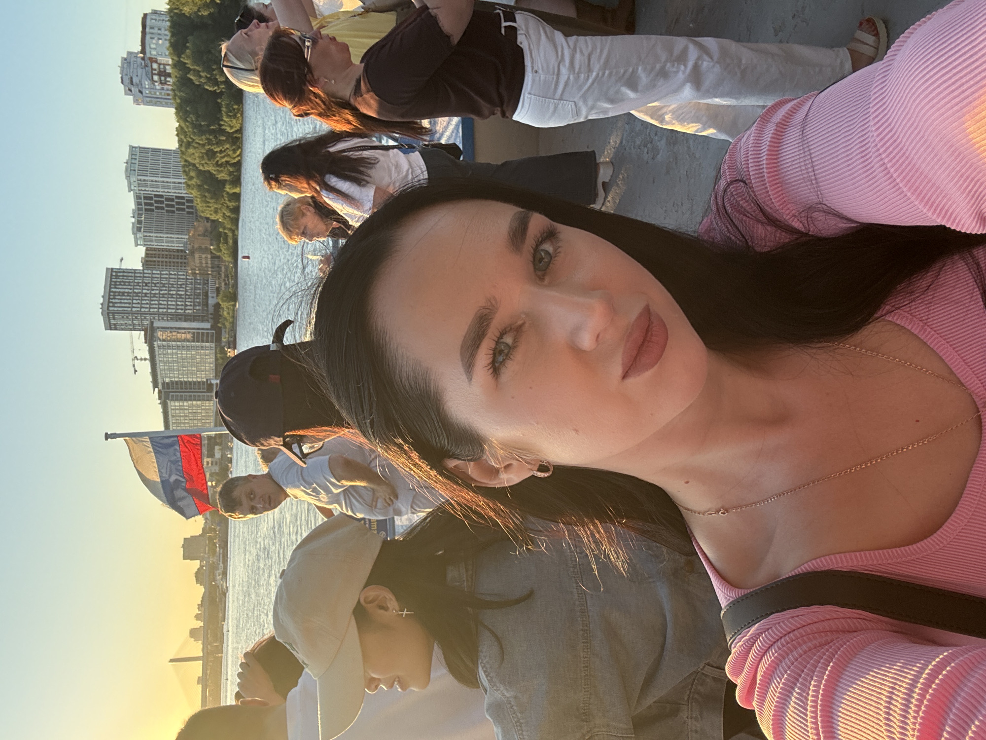 A group of people are on a boat on the Ob River in Novosibirsk, Russia, during sunset. The main subject, a woman with long dark hair and a pink top, is taking a selfie. Behind her, people are standing and looking around, with a Russian flag flying in the background. The cityscape of Novosibirsk is visible on the horizon, with tall buildings and trees lining the riverbank. The setting sun casts a warm glow over the scene.