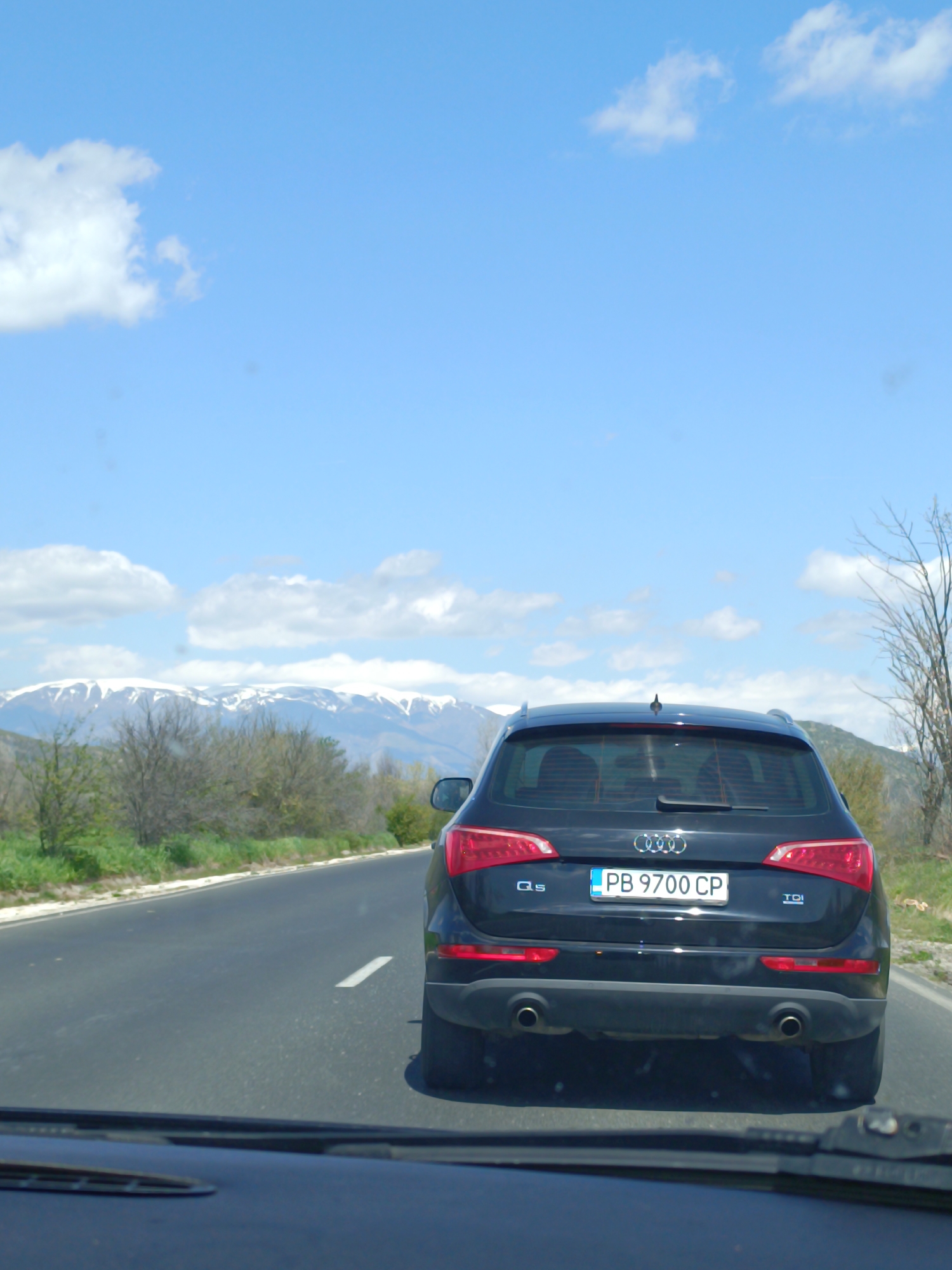 The image shows a black Audi Q5 driving on a road with a white line marking the lane. The car has a Bulgarian license plate "PB 9700 CP". In the background, there is a landscape with mountains partially covered in snow under a blue sky with some clouds. The shot is taken from inside a vehicle. The city and country are indeterminate.