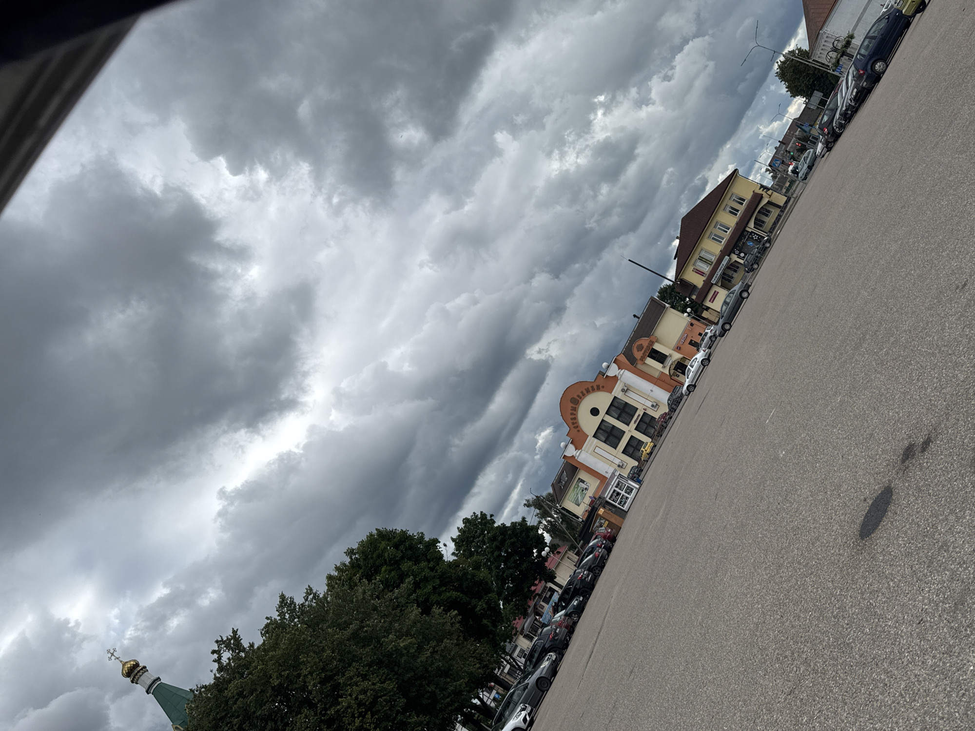 The image depicts a parking lot in the city of Maladzyechna, Belarus, under a heavily clouded sky. On the left, a green-spired building with a golden dome and a cross is visible. Adjacent to it are lush green trees. In the background, several buildings line the street, including a multi-story structure with an orange facade and arched windows, and a yellow building with a red roof. A row of parked cars, including a white sedan, several dark-colored vehicles, and a blue hatchback, occupies the parking lot. The ground is paved with asphalt, and there are some dark patches on its surface. The overall scene suggests an overcast day.