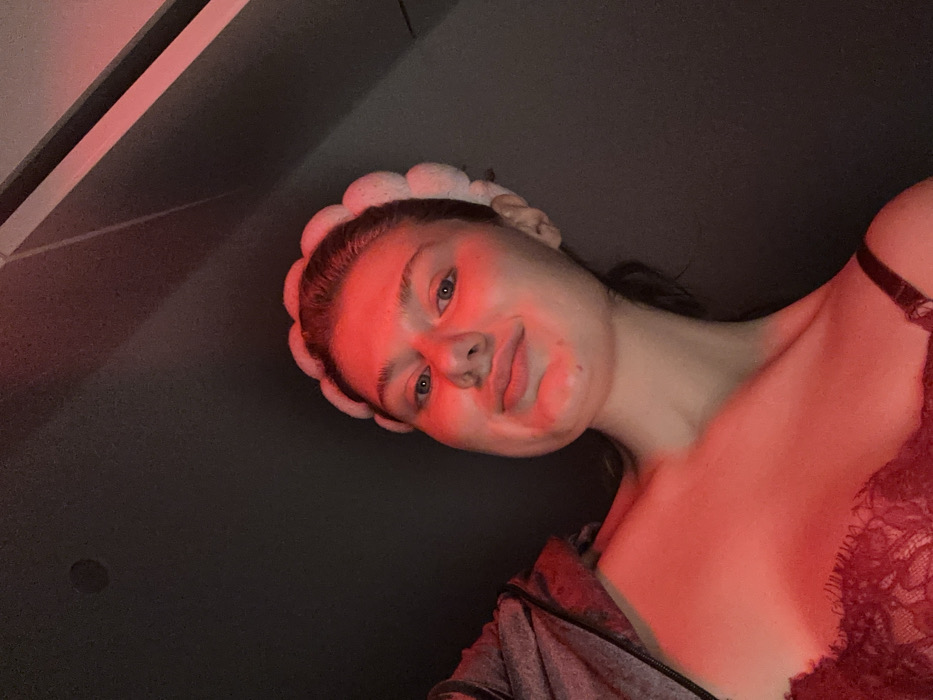 A woman's face is the focal point, illuminated by red lighting. She wears a soft, padded headband and has clear skin, save for a few spots on her cheek. Her eyes are blue and she looks directly at the camera. The shot is taken from a low angle, revealing part of her chest and a gray jacket or top with red lace. The background is a dark, solid color.