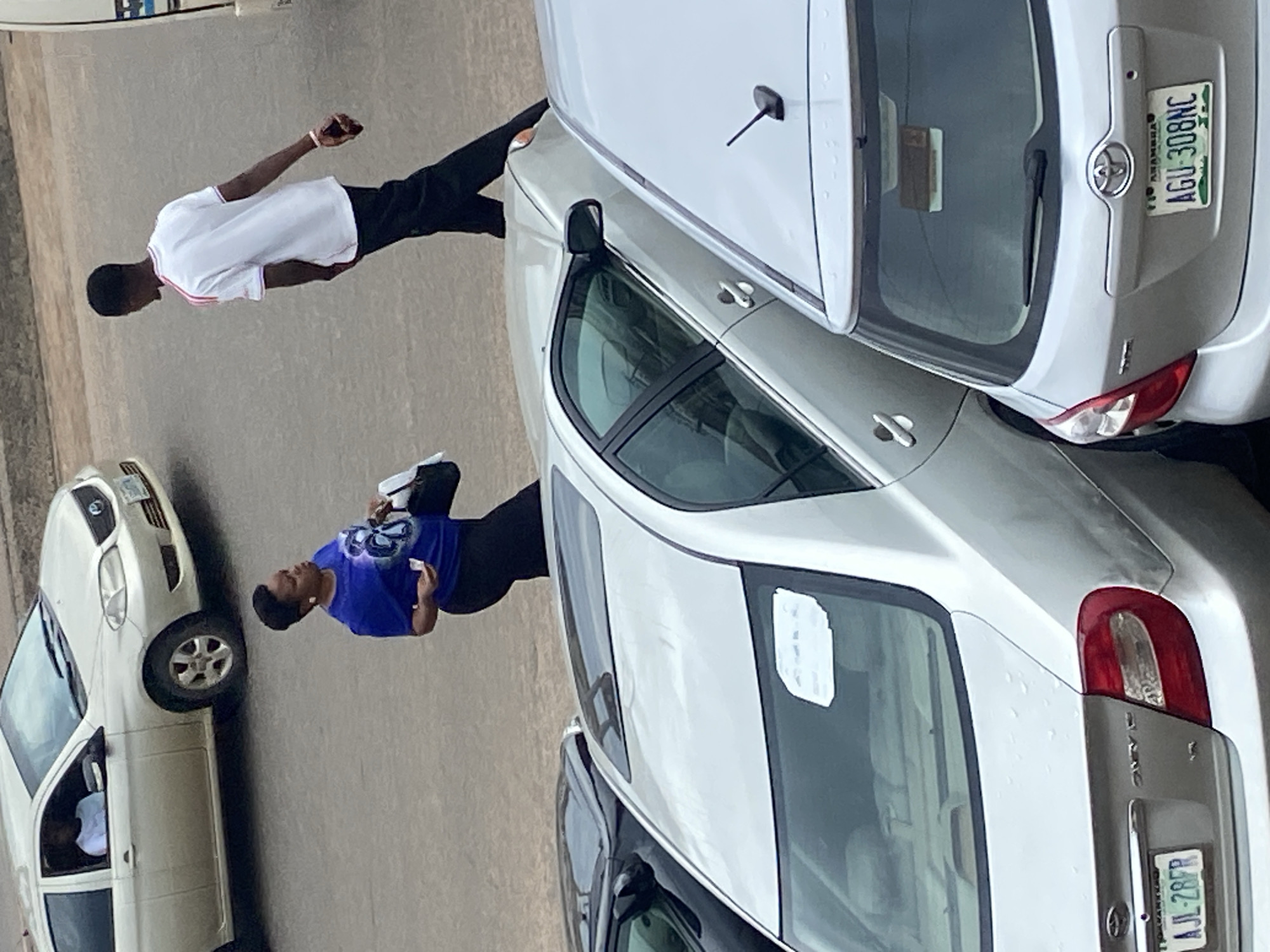 The image shows two people and several cars on a paved surface, possibly a street or parking lot, in Ogbunike, Nigeria.
The first person, a young man, is wearing a white t-shirt with red trim on the collar and sleeves, and dark pants. He is standing with his left arm outstretched, and appears to be looking towards his left.
The second person, a woman, is wearing a blue t-shirt with a graphic design and dark pants. She is holding a white object in her hand and looking down. She is standing next to a white car.
Several cars are visible in the image. One is a silver sedan, with its rear end facing the viewer. Another is a white sedan, which appears to be parked. A third car, mostly obscured, is a dark color and is parked behind the silver sedan. The license plate of the silver sedan is visible and reads "AGU-308NC". The license plate of the third car reads "AJL-28FR".
The scene appears to be set during the daytime. There are no clear indications of weather. The environment seems to be urban or suburban, consistent with a Nigerian location. The overall mood is neutral.