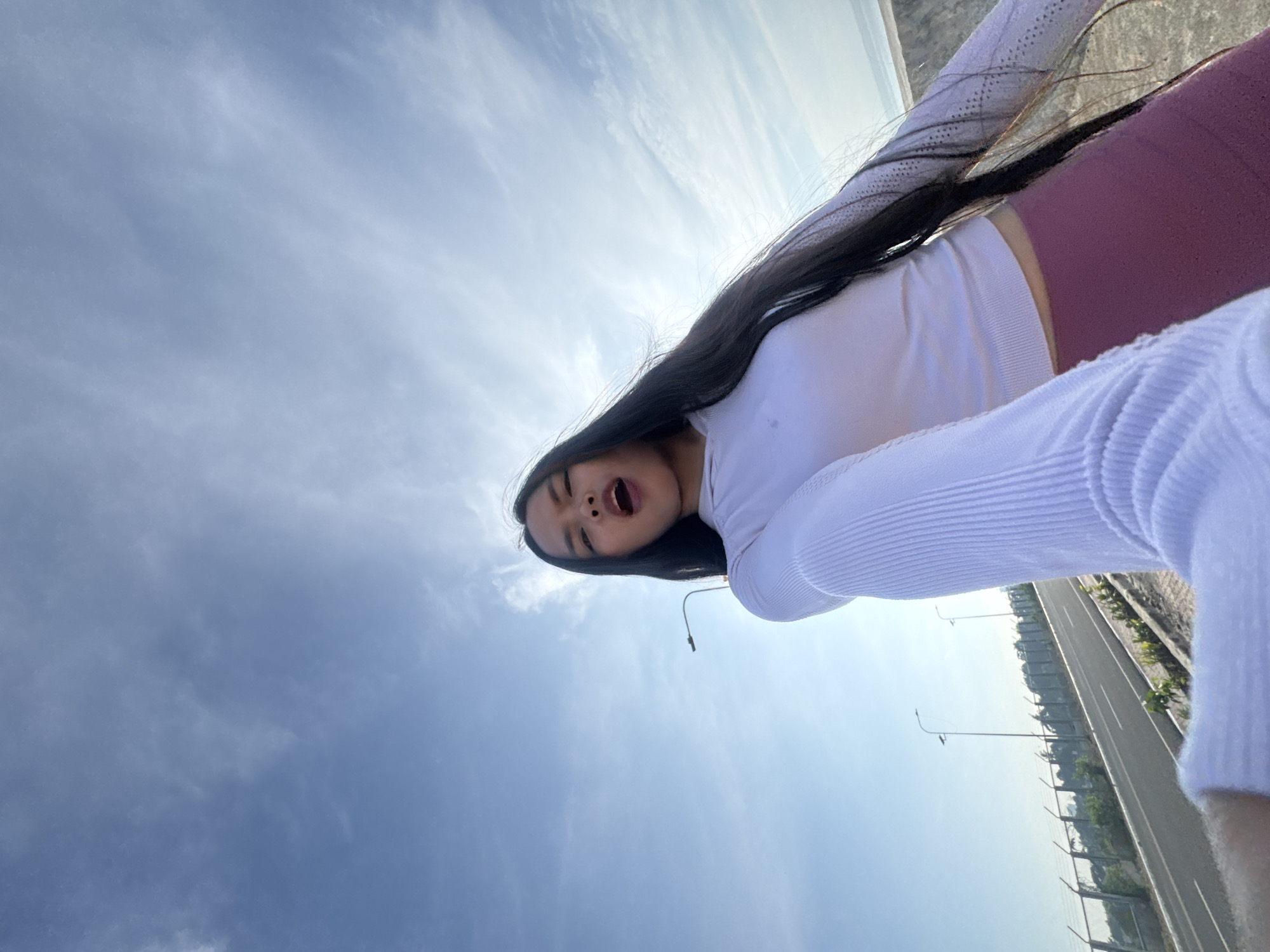 A young woman with long, dark hair is captured in a low-angle selfie outdoors against a bright blue sky with scattered clouds. She is wearing a white long-sleeved top and purple leggings, and her mouth is open as if speaking or laughing. The background features a road with streetlights, a metal fence, and some vegetation, suggesting an urban or semi-urban environment. The lighting indicates it is daytime, likely morning or afternoon. Without specific landmarks, pinpointing the exact location within Davao City is not possible from the visual information alone.