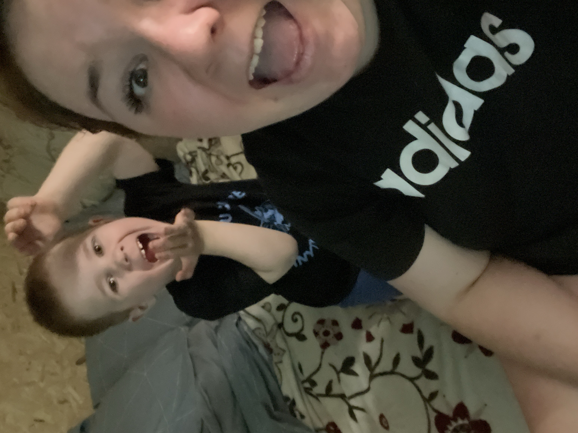 A selfie-style photo shows a person wearing a black Adidas t-shirt with their mouth open, positioned above and to the right of a young child. The child is lying on what appears to be a bed with floral patterned bedding. The child is also smiling and seems to be interacting with the camera. A gray or blue blanket is also visible. The location is unknown; there is no definitive evidence to indicate it is Chaikovsky, Russia.
