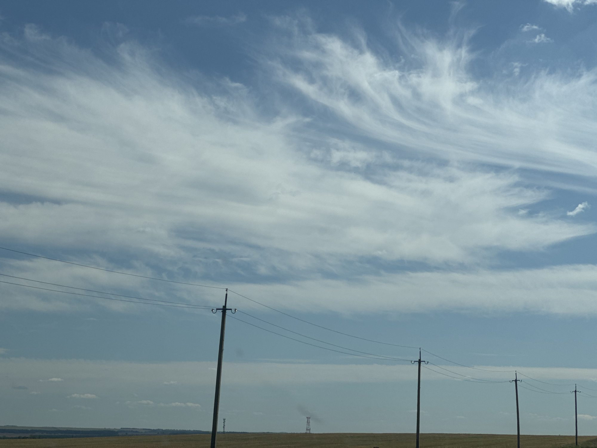 The image depicts a wide, flat landscape under a blue sky with wispy white clouds. In the foreground, several utility poles with wires stretch across the frame from left to right. The landscape itself appears to be a field of dry, golden grass or crops, with a distant tree line on the horizon. There are no people visible in the image. The overall scene is one of a rural, possibly agricultural, setting. The specific location in Klenovyi, Russia cannot be determined from the visual information alone.