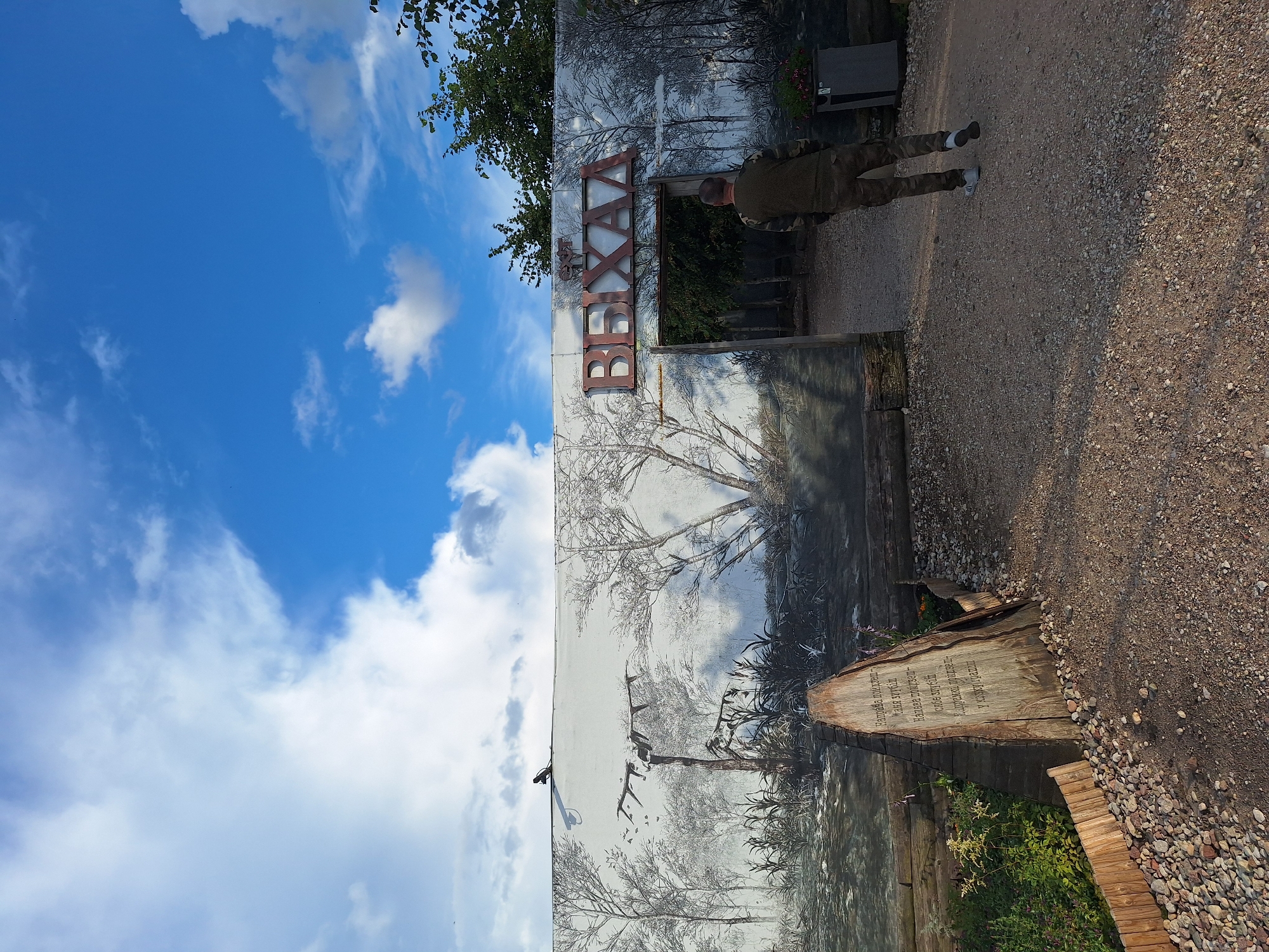 In the city of Малахавецкі сельскі Савет, Belarus, a man in camouflage clothing walks away from the camera, entering a gated area with trees visible beyond. He is positioned on a gravel pathway in front of a large white wall adorned with a mural depicting trees and birds. The mural is in a monochromatic, silhouetted style. Above the gated entrance, large metallic letters spell out "ВЕХАА". To the left of the entrance, a triangular wooden sign with text carved into it stands in front of the mural, accompanied by a low wooden fence and plants. The sky above is a bright blue with scattered white clouds.