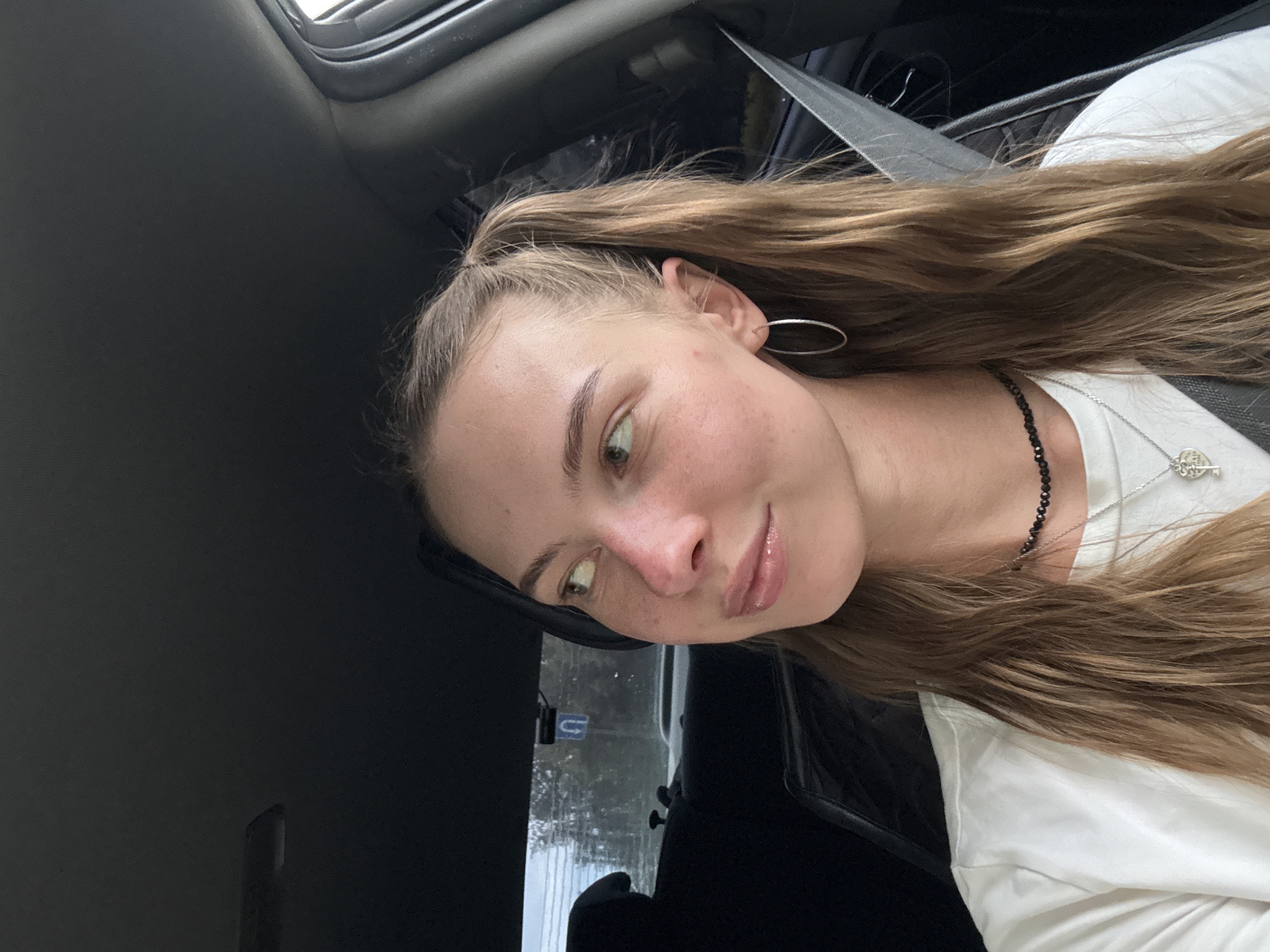 A young woman is pictured inside a car, looking to her left. She has light skin, brown hair styled in pigtails, and is wearing a white t-shirt, a black beaded necklace, and a silver necklace with a key pendant. She also has hoop earrings. The background shows the interior of a car, including a seatbelt, and through the windows, trees and a street sign are visible. The street sign appears to be a blue sign with a white arrow indicating a U-turn, consistent with road signage found in Perm, Russia.