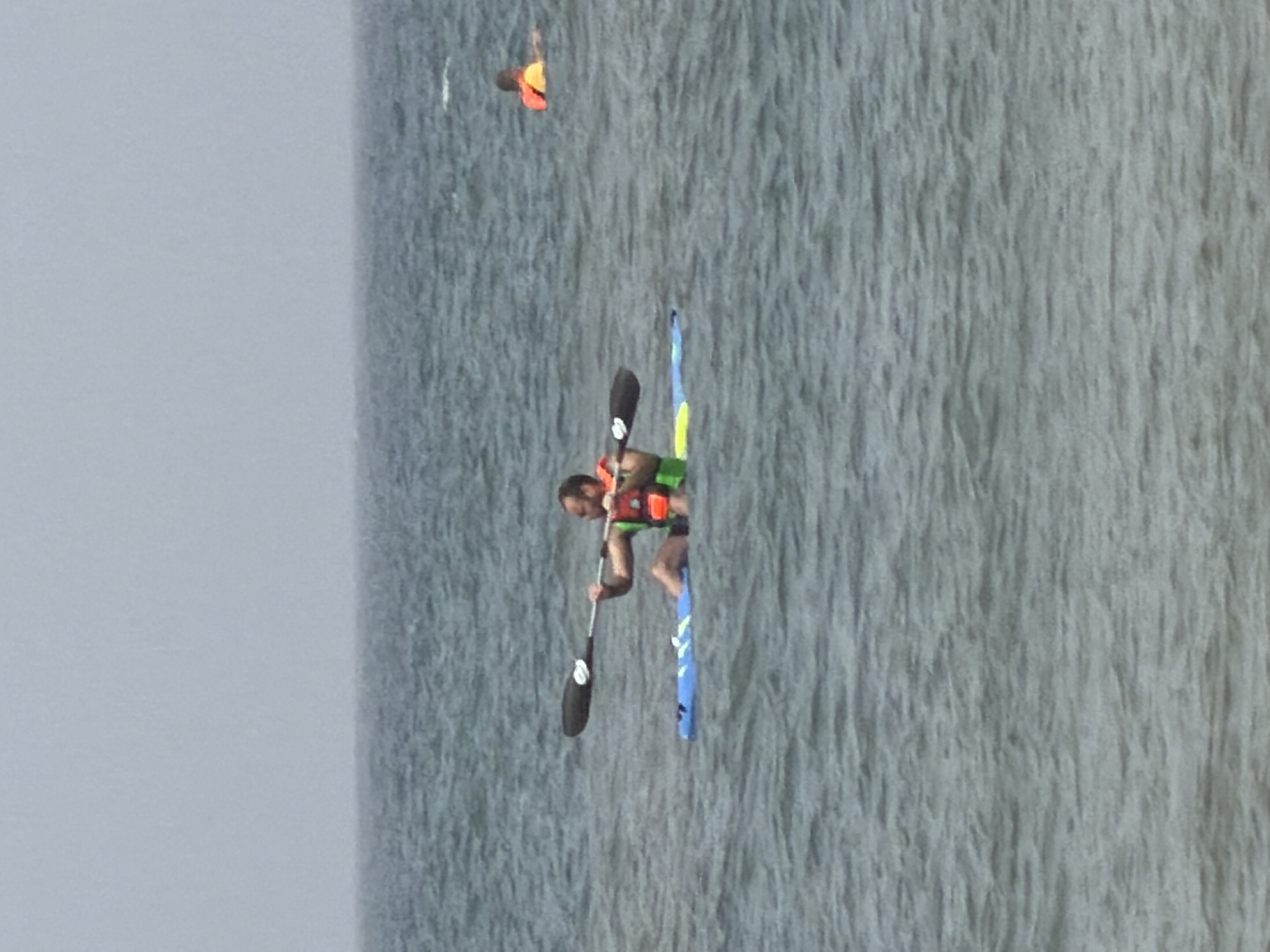A man wearing a life jacket is kayaking on the sea, holding a paddle. A second person wearing a similar life vest is visible in the distance. The scene appears to be taking place in the sea. No information in the image confirms this location to be Tel-Aviv.
