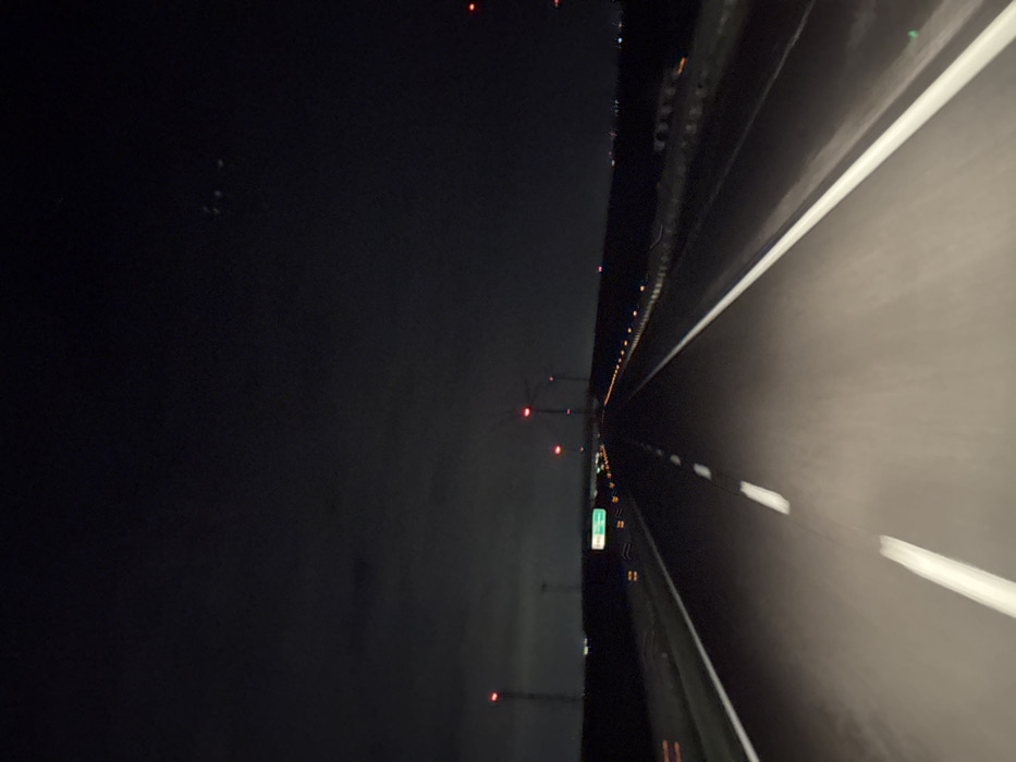 The image depicts a nighttime scene on a multi-lane highway within the area of Orta Nova, Italy, viewed from a vehicle in motion. The road surface, illuminated by the viewpoint vehicle's headlights, shows clear white dashed lane markings in the center and a solid white line defining the right edge of the immediate lane. To the left, a green informational road sign with illegible white text is visible in the middle distance. The horizon features numerous tall structures, identifiable as wind turbines, on both sides of the highway, each equipped with a prominent red obstruction light. Along the right side of the highway, a metal guardrail runs parallel to a line of smaller, amber-colored lights extending into the distance. The sky above is dark with minimal, faint cloud cover. No individuals, specific vehicles other than the implied viewpoint, or explicit actions are discernible.