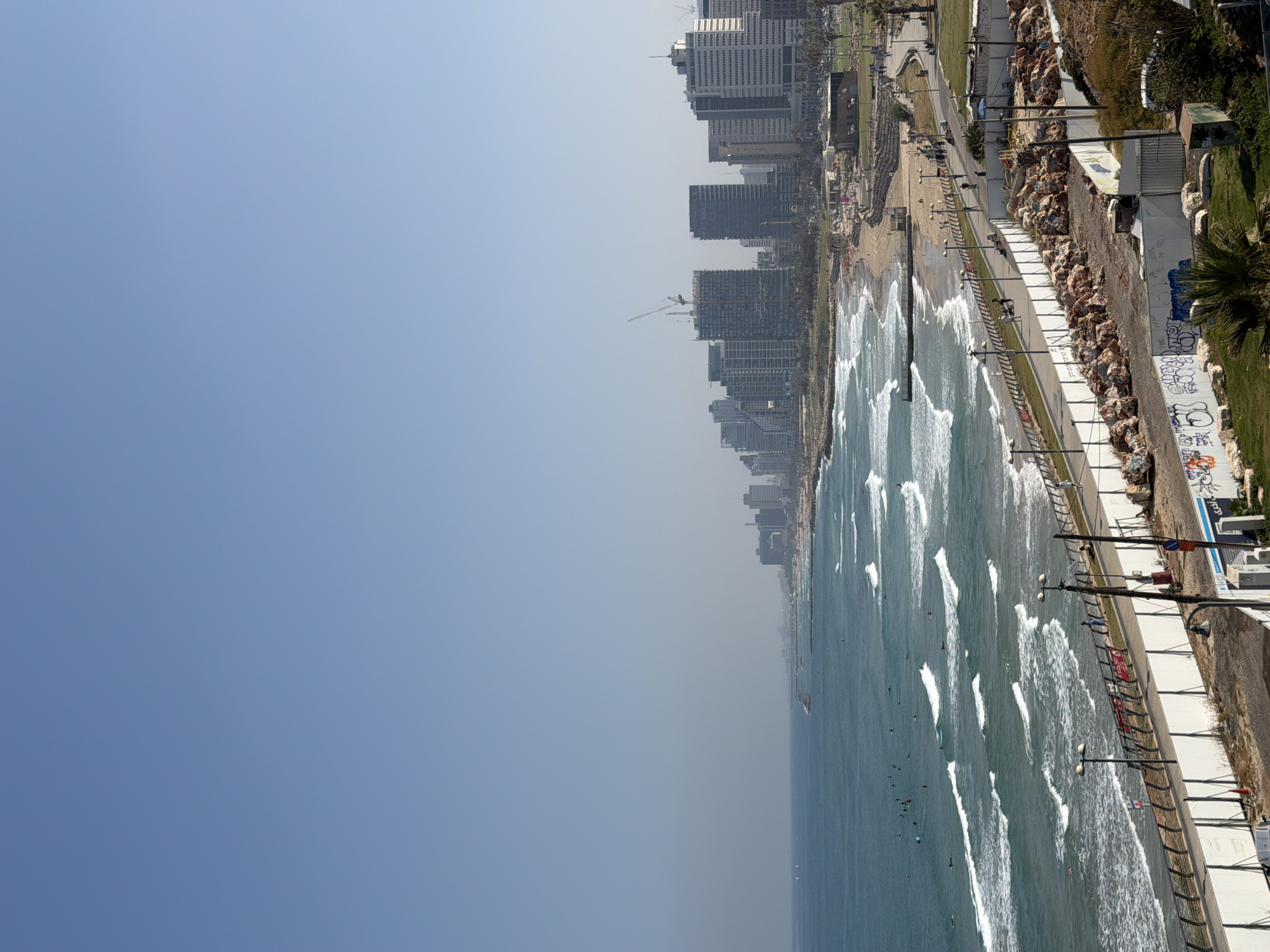 The image shows a view of the Tel Aviv coastline in Israel. The Mediterranean Sea is on the right, with waves breaking along the shoreline. Numerous surfers are visible in the water. A beach runs along the coastline, adjacent to a promenade with lampposts and a low barrier. Behind the promenade, there's a raised area with rocks, vegetation, and a concrete wall, which has graffiti. The city skyline, including several high-rise buildings and construction cranes, is visible in the top left corner. The sky is clear and light blue.