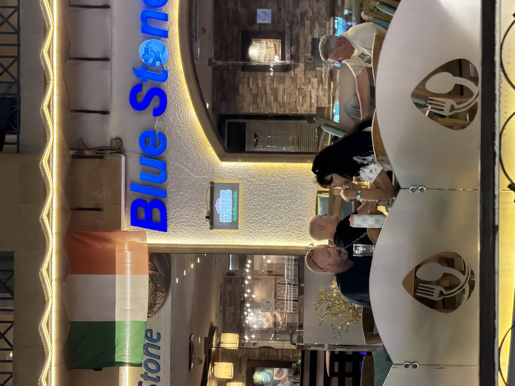 The image depicts an outdoor seating area of a restaurant in Sharm El Sheikh, Egypt. In the foreground, a decorative metal partition with a cutout of a fork and spoon is visible. Four people are seated at tables. The person on the far left is a bald white male wearing a dark t-shirt, looking down and holding a glass. Next to him, partially obscured, is another bald white male with his head turned to the right. In the center, a woman with long dark hair and a white floral top is seated, looking towards the right. To her right, a white male wearing a light-colored t-shirt is seated, looking to the left. Behind the diners, the facade of a building features illuminated signage that reads "Blue Stone." An Irish flag is displayed above the signage. A sign for "DESPERADOS Tequila Flavoured Beer" is also visible on the building. The setting appears to be evening, with warm lighting illuminating the scene.