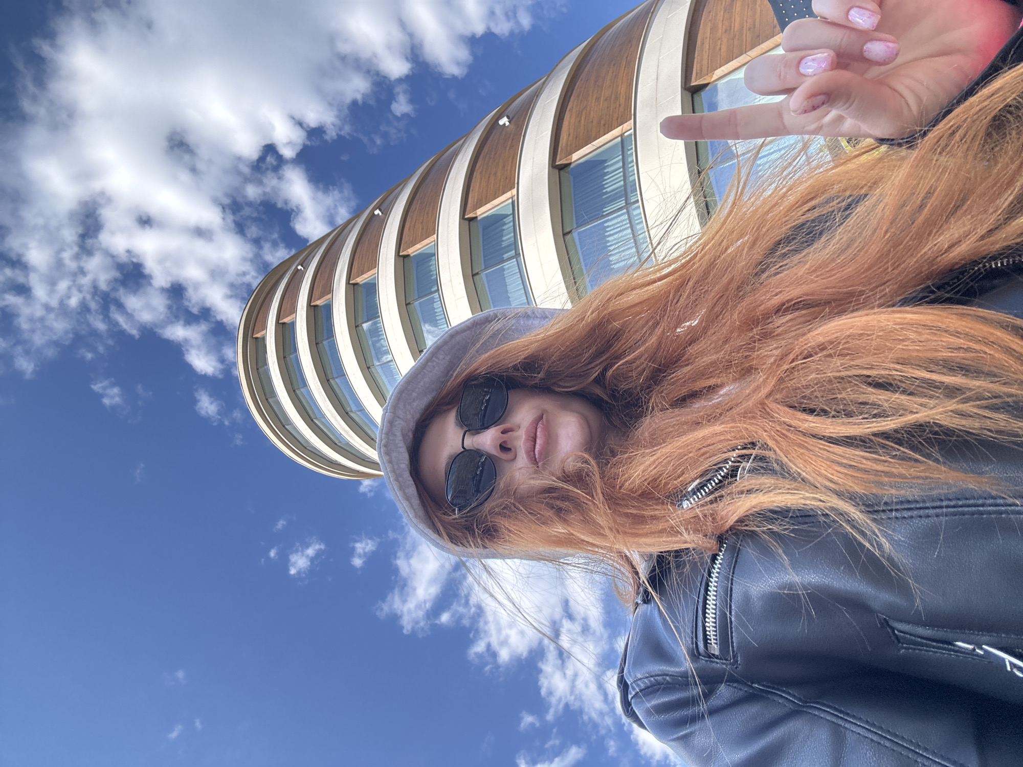The image shows a woman standing in front of a building under a partly cloudy, bright blue sky. She is wearing a hooded sweatshirt, sunglasses, and a black leather jacket. Her hair is long and reddish-orange. She is holding up her middle finger. Behind her is a cylindrical building with light-colored bands and rectangular windows reflecting the sky. Based on the architecture and context clues, the location is likely in Budva, Montenegro.