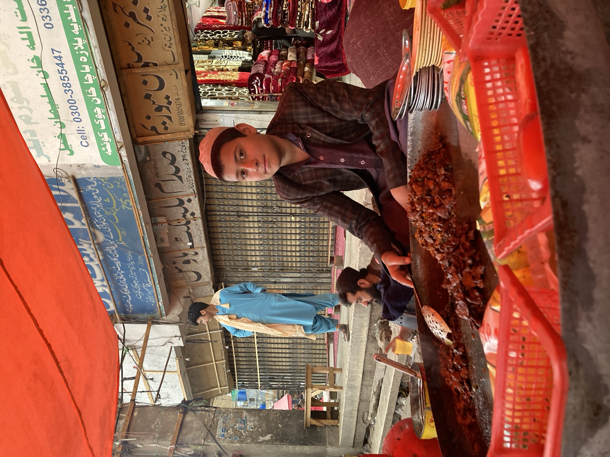 The image depicts an active street food stall operating under a large, bright orange tarp in Quetta City Tehsil, Pakistan, specifically near Baldia Plaza Bacha Khan Chowk.
In the immediate foreground, a young male, appearing to be a teenager, sits behind a large, dark metal pan (tawa/karahi) filled with dark brown, cooked meat pieces and vegetables. He wears a light pink kufi, a patterned brown blazer over a maroon collared shirt, and maroon trousers, looking directly at the camera with a neutral expression. His right hand rests near a stack of small, silver-colored metal plates and yellow plastic plates. Several red plastic baskets are visible in front of the pan, one containing red objects, possibly tomatoes. A metal spatula with perforations is resting within the cooked food.
To the left of the main subject, a bearded adult male in dark clothing is seated on the ground, leaning forward. He holds a small, clear cup containing a yellow liquid in his left hand, while his right hand is positioned close to the cooking pan.
In the mid-ground, on a raised concrete platform to the left, a young adult male stands with his hands in his pockets. He is dressed in a light blue shalwar kameez with a beige scarf draped over his left shoulder, looking towards the left of the frame. A small, light-colored wooden stool or table is situated near his feet.
The background features several commercial buildings. One prominent blue sign above the main scene, written in Urdu/Pashto, clearly identifies "بالدیہ پلازہ باچا خان چوک کوئٹہ" (Baldia Plaza Bacha Khan Chowk Quetta) and includes a cell number (0300-3855447). Another sign reads "سلطان میوزیم" (Sultan Museum). Most shops in the background are secured with closed metal roller shutters. To the far right, inside an open shop doorway, numerous colorful blankets and quilts are stacked high, and a partial view of another person is visible within the shop. The ground level is uneven, displaying typical street detritus.