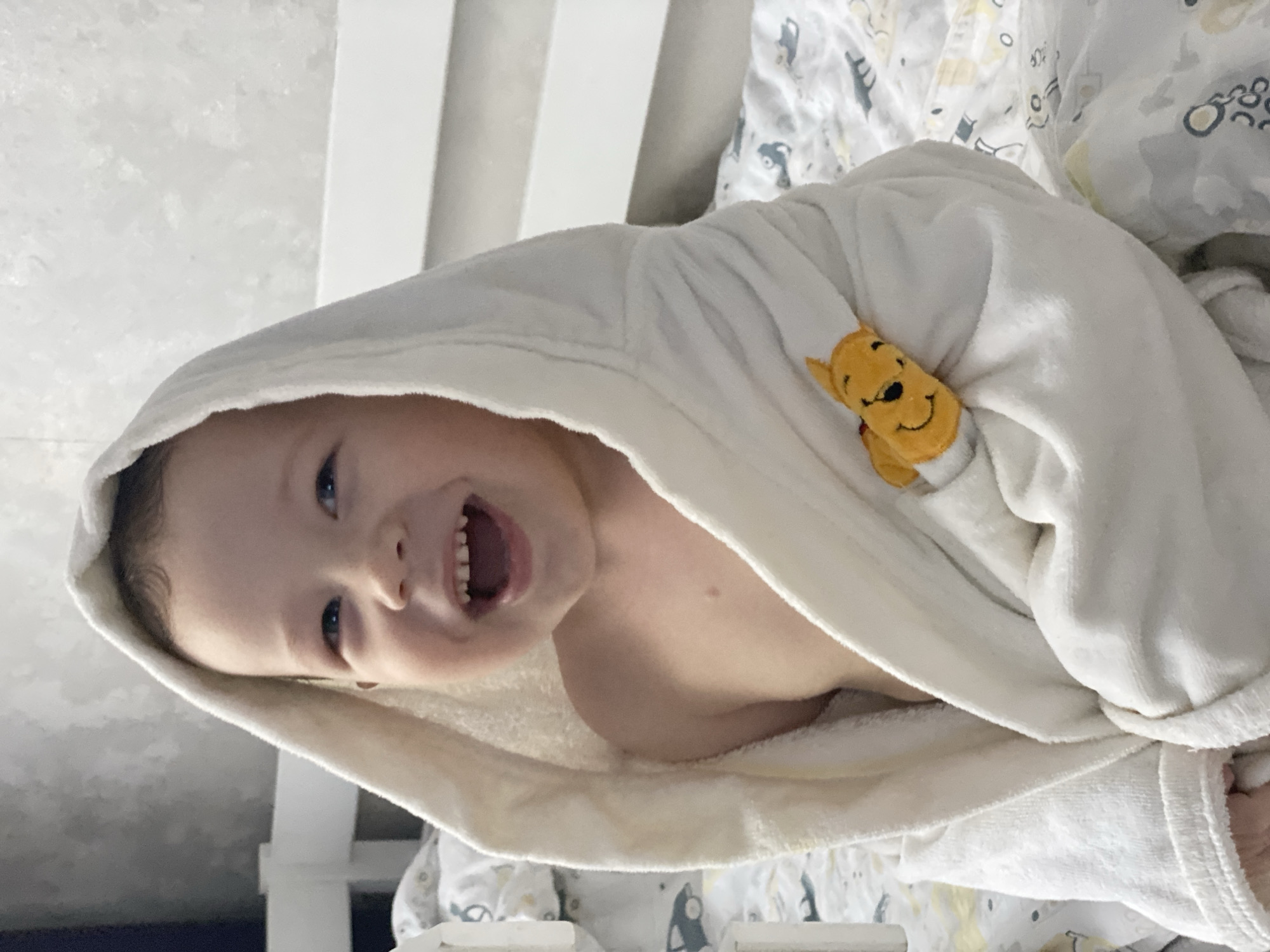 A young child, likely a toddler, is captured in a joyful, close-up portrait, looking directly at the viewer with a wide, open-mouthed smile, revealing small teeth. The child has light skin, bright eyes, and appears to have dark hair visible from under a hood. They are wearing a soft, white hooded bathrobe, possibly made of terry cloth, which covers their body and shoulders. A small, yellow Winnie the Pooh character, appearing as an appliqué or plush element, is attached to the right side of the bathrobe, adding a whimsical touch.
The background primarily shows a plain, light-colored surface, possibly a white wall or the headboard of a bed, on the left side of the frame. To the right and below the child, patterned bedding is visible, featuring a white base with light gray and yellow motifs, possibly depicting cars or other playful shapes, suggesting a child's room or bed. The child's expression conveys happiness and playfulness, possibly during a moment of laughter or just after a bath. The scene is indoors. No text is visible in the image. The location context provided is Sochi, Russia.