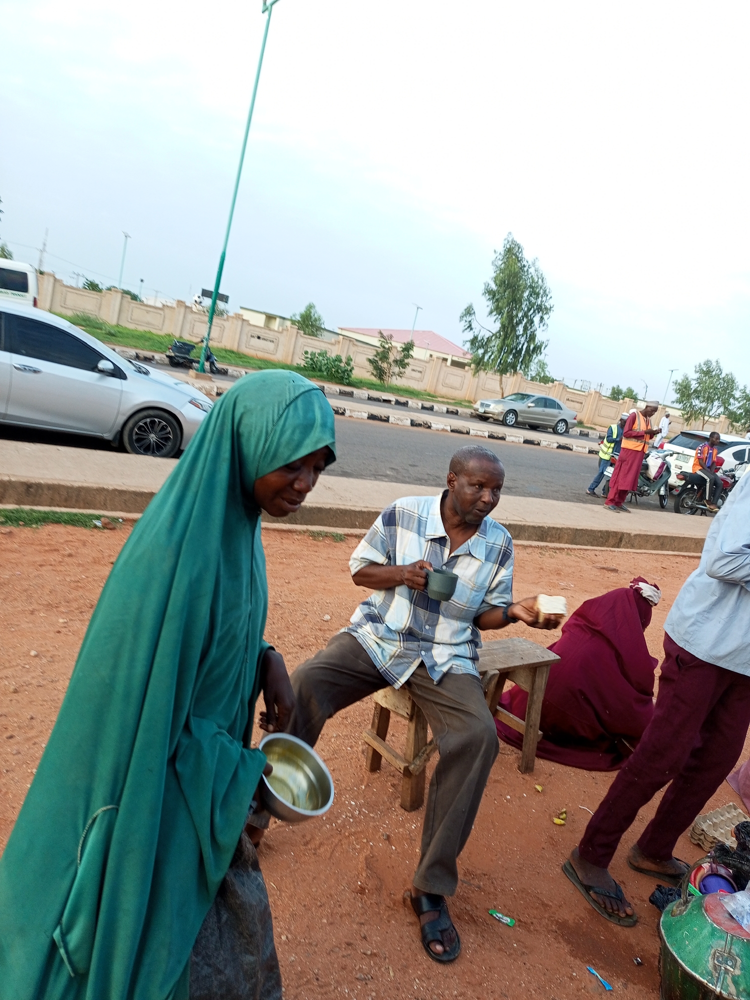 A medium shot shows a group of people near a street in an outdoor setting, likely a city. In the foreground, a woman in a teal hijab and dark garment holds a metallic bowl. To her right, a man in a blue and white checkered shirt and brown pants sits on a wooden stool, holding a dark mug and a piece of white bread. Behind them, a person in a maroon garment is seated. Further in the background, several individuals are visible near motorcycles and vehicles on a paved road with a decorative wall and trees. The ground is a reddish-brown, dusty surface. The sky is overcast.