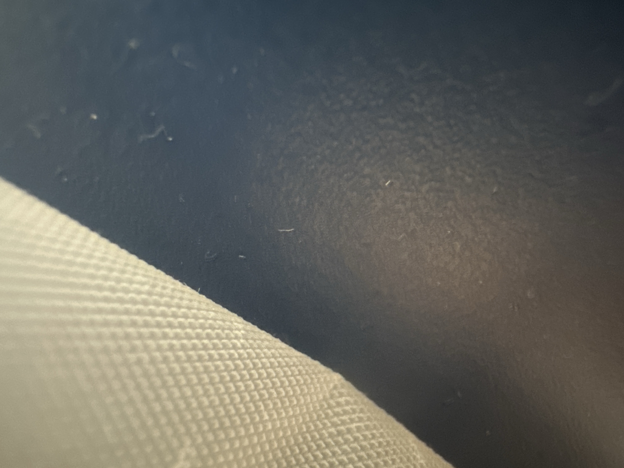 The media file is a close-up, low-angle shot of a textured, white object in the foreground, which appears to be a fabric or surface with a repeating pattern of small, raised bumps. This object occupies the bottom left portion of the frame and extends diagonally upwards and to the right. In the background, a dark, textured surface is visible, predominantly a deep blue or black color. This background surface has a slightly mottled or grainy appearance.
Scattered across both the foreground object and the background are small, white specks, which are likely dust or debris. There are no people visible in the image. The lighting suggests an indoor environment, possibly with artificial light, as there are no strong shadows or indications of natural light sources like sunlight. The image is underexposed in some areas, particularly the background, which creates a sense of depth and mystery. The focus is shallow, with the foreground object being sharper than the background. No text is visible in the media file. The overall impression is abstract, focusing on texture and pattern rather than a discernible scene or activity. The location context of Belgrade, Serbia, does not provide any specific visual cues within the image itself.