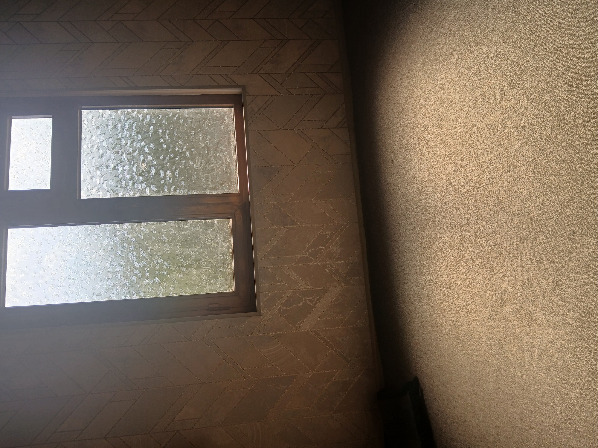 The image shows an interior room with patterned wallpaper and a carpeted floor. A wooden window with textured glass is visible on the back wall. The lighting suggests it is daytime, with sunlight streaming through the window and creating a bright spot on the floor. No people or specific actions are depicted. The scene appears to be a typical domestic interior. The location of Kushkayragach, Uzbekistan, is not discernible from the visual information presented in the image.