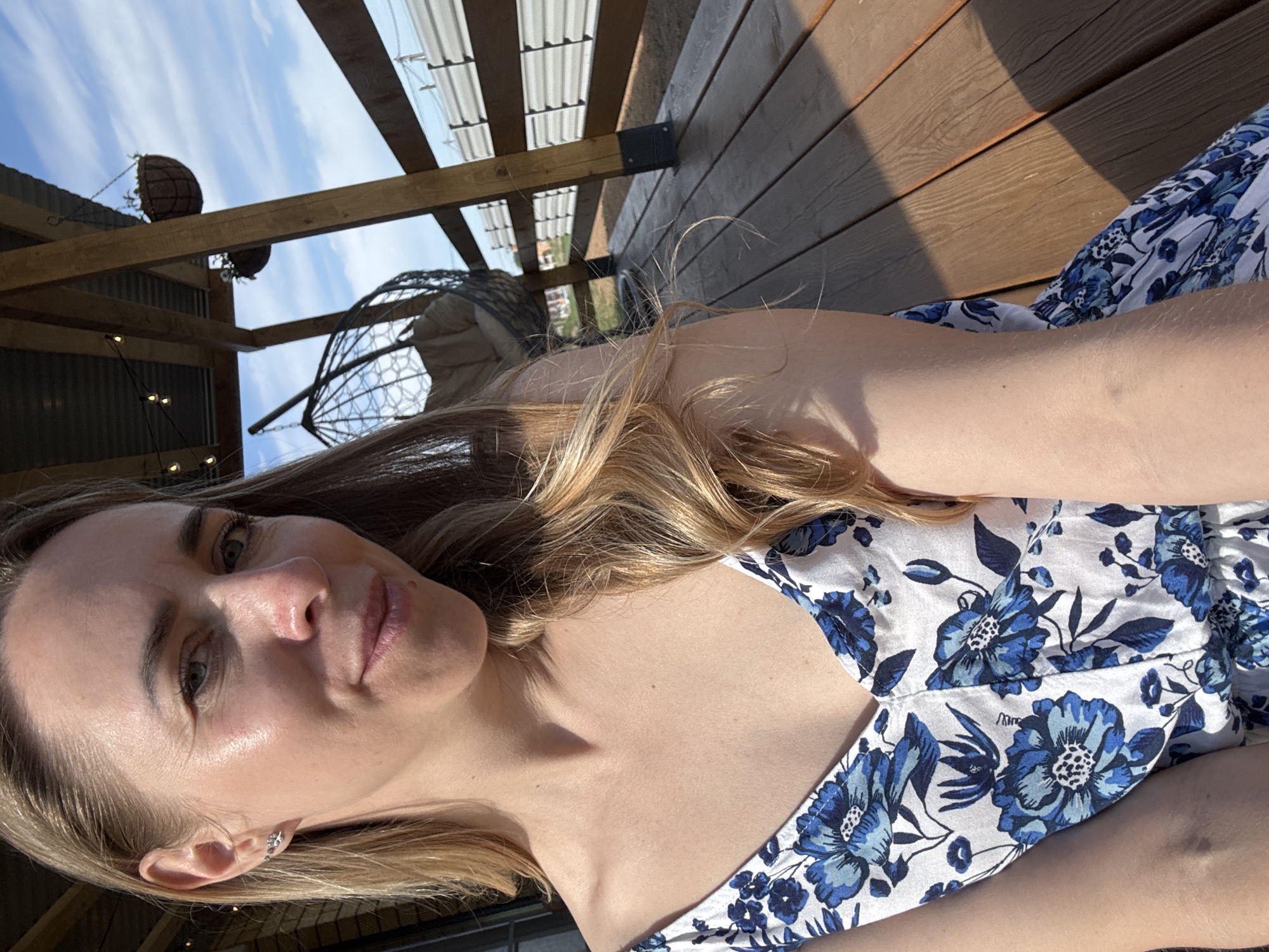 A young woman with shoulder-length blonde hair is posing for a selfie outside on a sunny day. She is wearing a white top with blue floral patterns. Behind her is a wooden deck with sunlight casting shadows on the planks. There is an open-air structure overhead with hanging planters and a wire chair partially visible. The location is possibly somewhere in Chaikovsky, Russia.