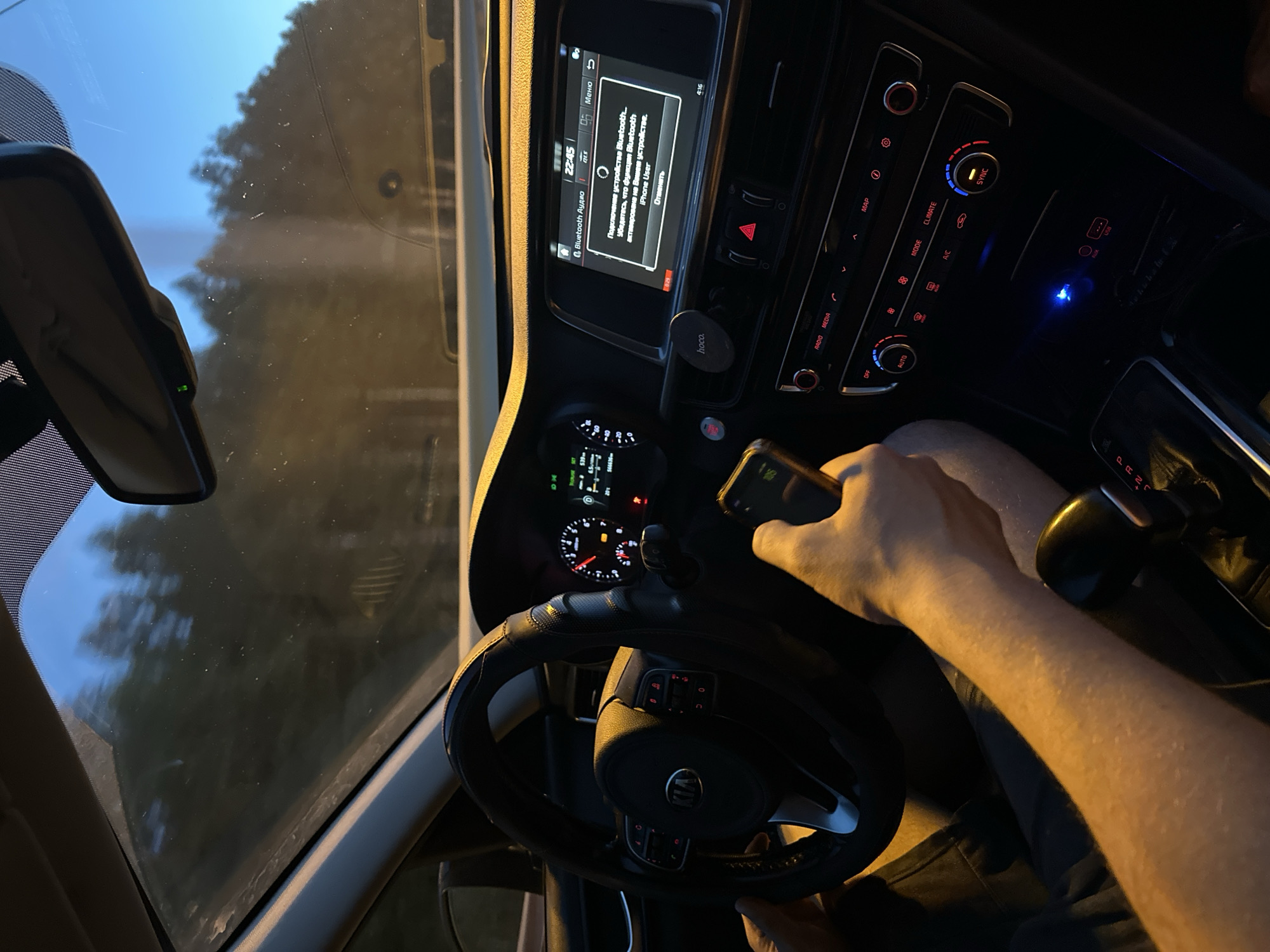 This image captures the interior of a car at dusk, with the scene oriented vertically. The driver's perspective is shown, with a hand holding a smartphone visible in the foreground on the right. The steering wheel, covered in black material, is prominent, and the car's dashboard features a digital display showing "22:45" and a Bluetooth connection prompt in Russian, indicating a connection attempt with an "iPhone User". The central console displays climate controls and media options. A blue LED light emanates from a USB port. Outside the car, seen through the windshield and side window, the sky is a twilight blue, and trees are silhouetted against it, suggesting a rural or suburban setting on the outskirts of Старые Решёты, Russia. The dim lighting and the activity of connecting a phone imply the person is either about to start a journey or has just arrived at their destination.