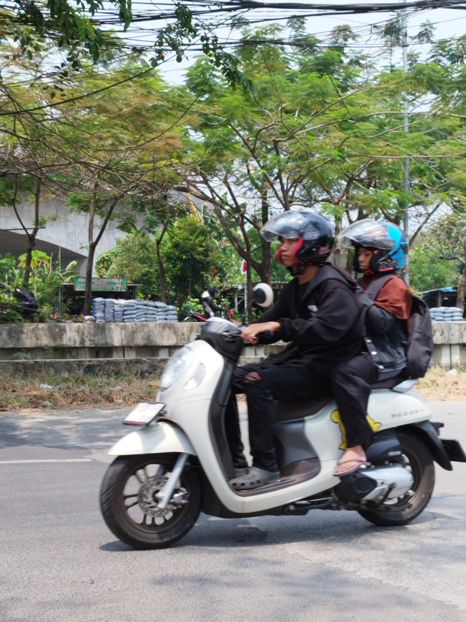 Two people ride a white scooter on a paved road in an urban environment. The person in front, driving the scooter, wears a black hoodie and black pants, and a black helmet with a clear visor. The person in the back, a child, wears a brown top, a denim vest, and a blue helmet with a clear visor. Both individuals are wearing helmets. The scooter has a license plate that is partially obscured. In the background, there is a concrete barrier, stacks of blue bags, trees, and overhead wires. The lighting suggests it is daytime.