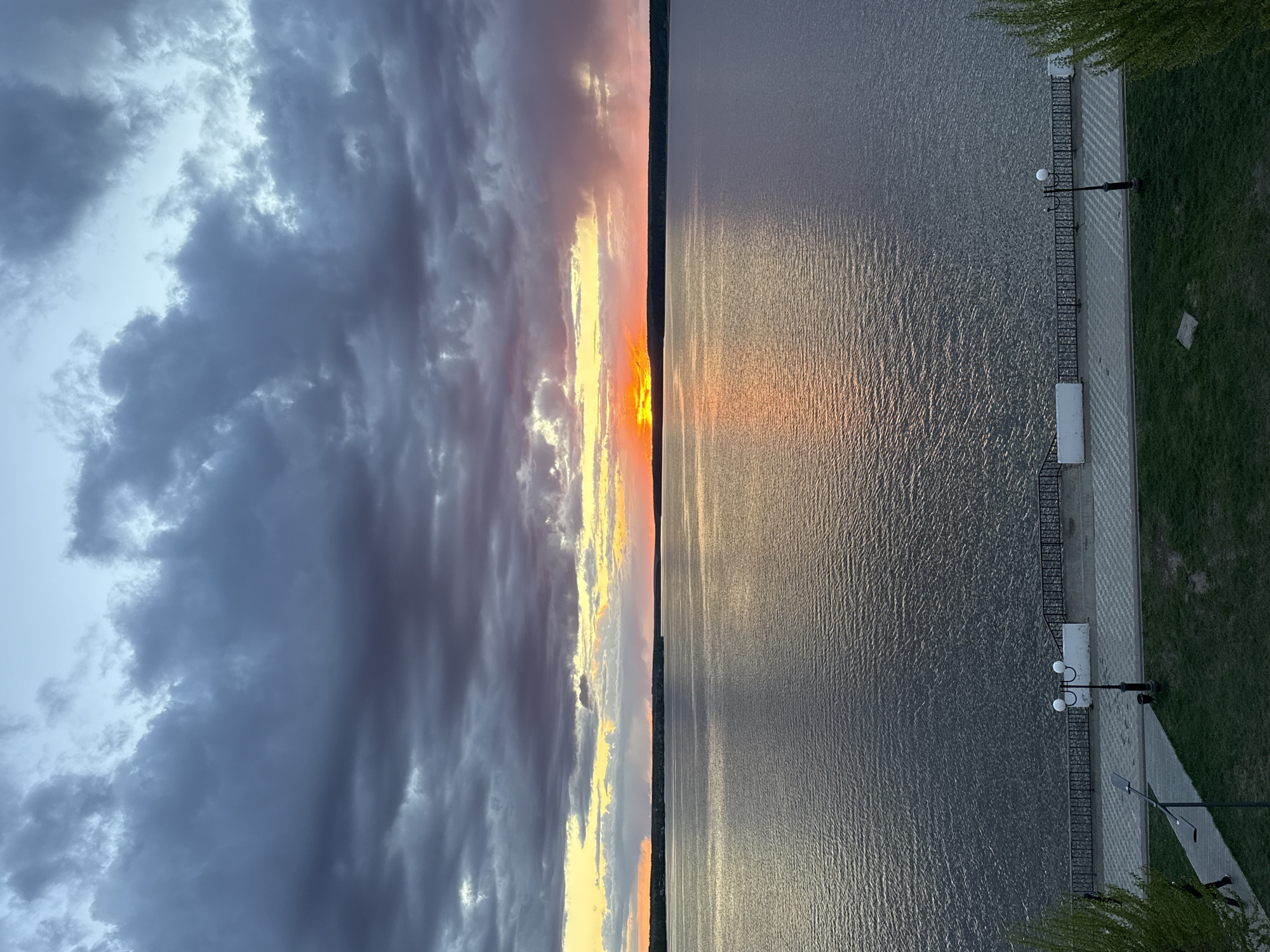 The image captures a sunset over a body of water, likely the Votkinsk Reservoir in Votkinsk, Russia, given the distinctive embankment visible. The sky features dramatic, dark clouds on the left transitioning to lighter, sunset-hued clouds in the center where the sun is setting. The body of water reflects the sunset colors, creating a mirrored effect. On the right side of the frame, an embankment with a patterned walkway is visible. Lamp posts and benches are situated along the embankment, and green grass borders the walkway. There are no people visible in the shot. The environment suggests a calm, tranquil evening.