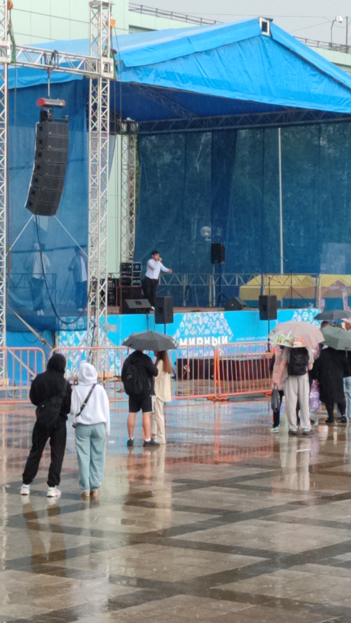 The image depicts an outdoor event in Mirny, Russia, under a blue canopy structure that covers a stage. A performer in a white shirt and dark pants is on stage, speaking into a microphone. Several speakers are visible on and around the stage. In front of the stage, a metal fence separates the performance area from a wet paved area where several people are gathered. Many of these individuals are holding umbrellas, indicating it is raining. The ground is reflective, suggesting puddles or recent rainfall. A banner with Russian text, possibly "Мирный" (Mirny), is visible on the stage backdrop. The overall scene suggests a public gathering or concert taking place despite inclement weather.