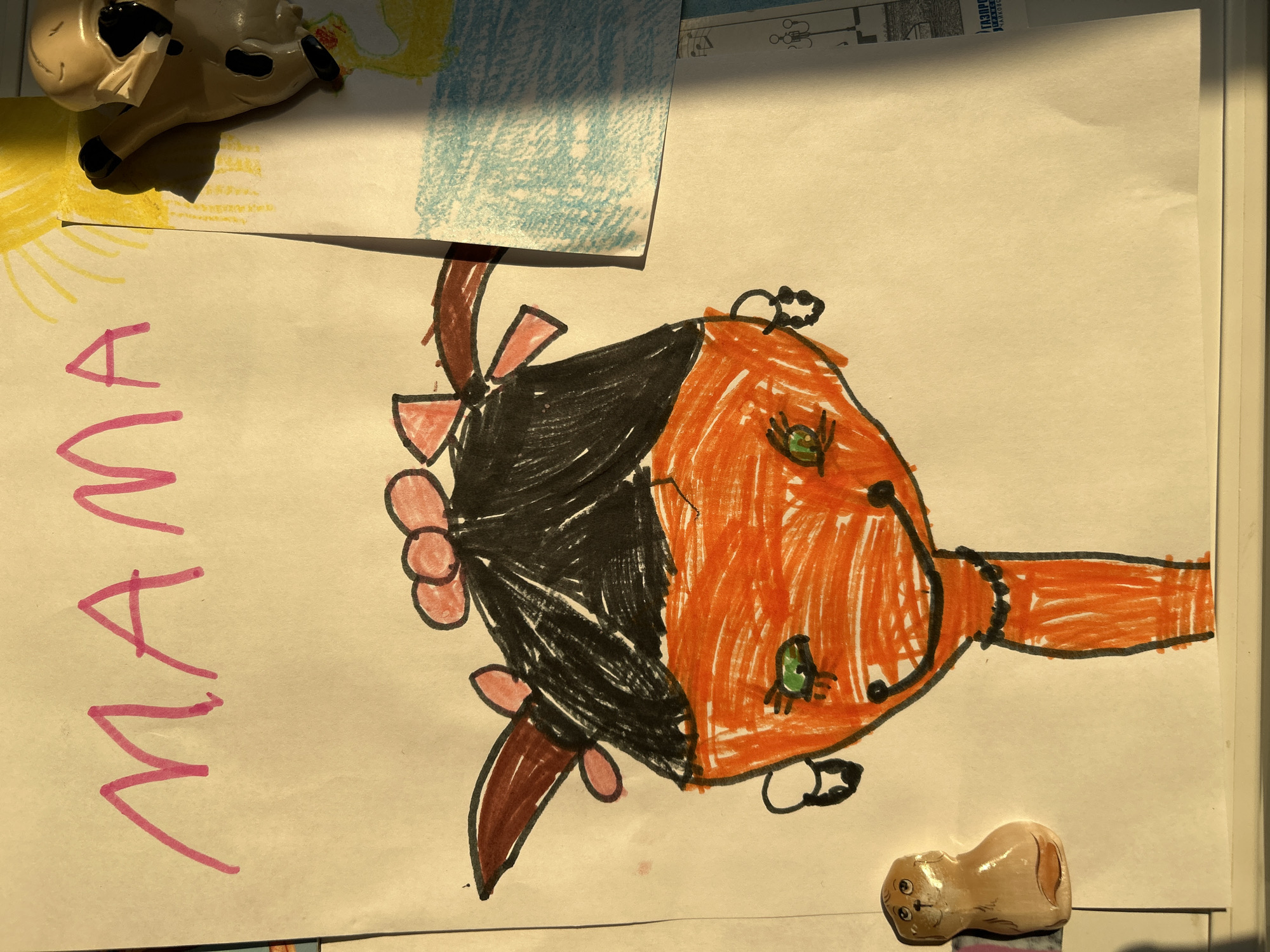 A child's drawing, oriented vertically, depicts a stylized face or head with vibrant colors. The drawing features an orange-colored face, black hair, and bright green eyes. Pink bows adorn the hair/head, complemented by a brown element, possibly a neck or shoulder, and a black beaded necklace. The word "MAMA" is written in pink block letters to the left of the drawing.
The artwork lies on a light-colored surface, likely a table or floor, and is partially covered by two other pieces of paper at the top left. One partially visible drawing above shows a yellow sun and a blue-colored area, suggesting another piece of child's art. A white and black cow figurine is positioned in the top left corner, resting on these papers. In the bottom right corner, a small, light brown human-like figurine with a striped base is also visible.
Strong, directional sunlight casts distinct shadows across the papers, indicating an indoor setting during daytime, possibly morning or late afternoon. In the background, beneath the main drawing, another paper or magazine sheet is visible with printed text and schematic diagrams. Fragmented text "ГАЗПРОМ" (GAZPROM) and "Чайковск" (Chaikovsk) can be discerned on this background paper, indicating the location as Chaikovsky, Russia. The overall scene evokes a domestic environment displaying a child's creative work.