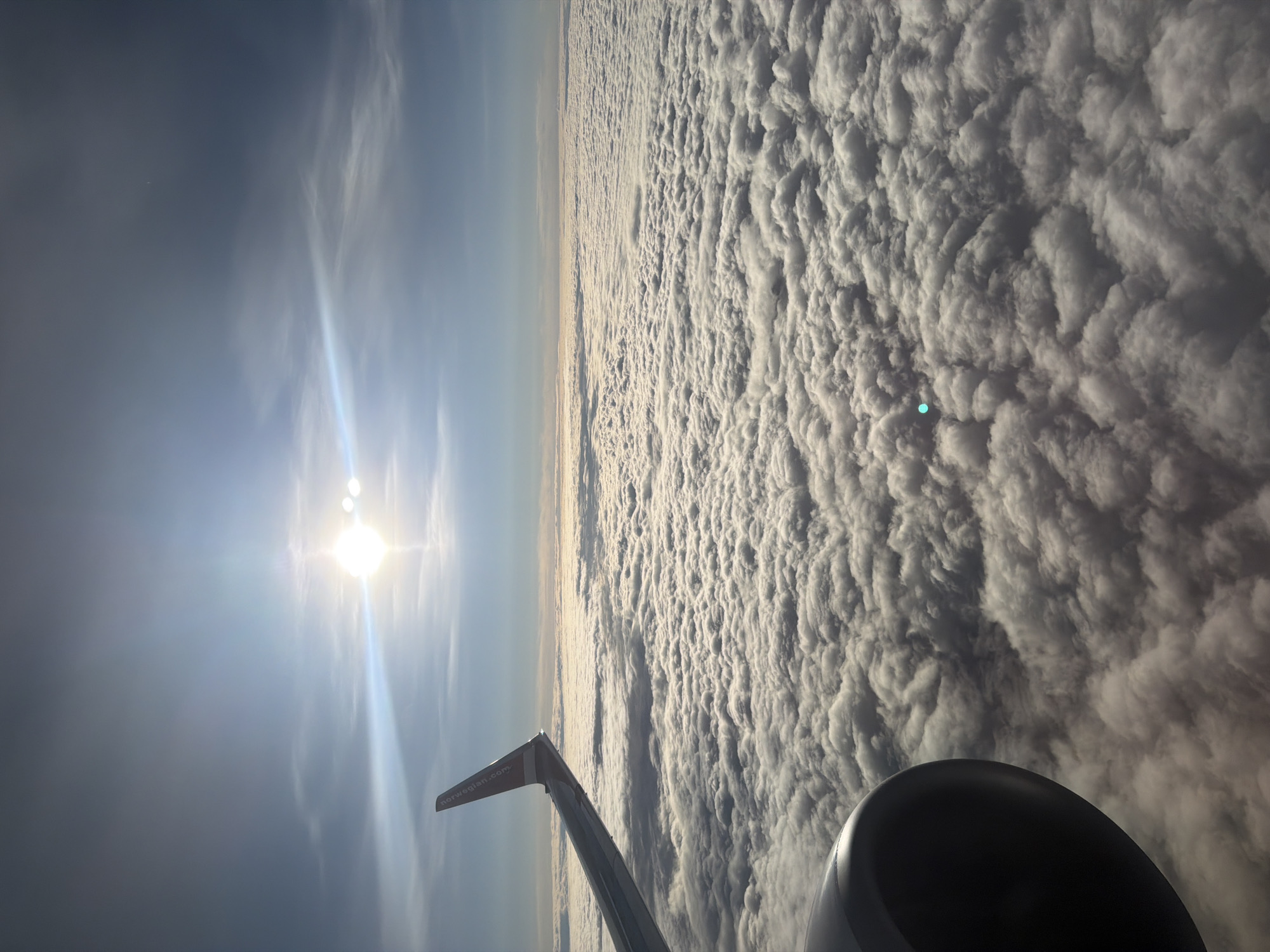 This is an aerial photograph taken from an airplane window, likely above Gardermoen, Norway. The sun is shining brightly in the upper portion of the frame, creating a lens flare effect. Below the sun, a vast expanse of white, fluffy clouds stretches to the horizon. A portion of an airplane wing, featuring the Norwegian logo, is visible in the upper left corner. In the lower left quadrant, a section of the airplane's engine is also visible, appearing dark and metallic. The overall scene depicts a flight over a cloud-covered landscape.
