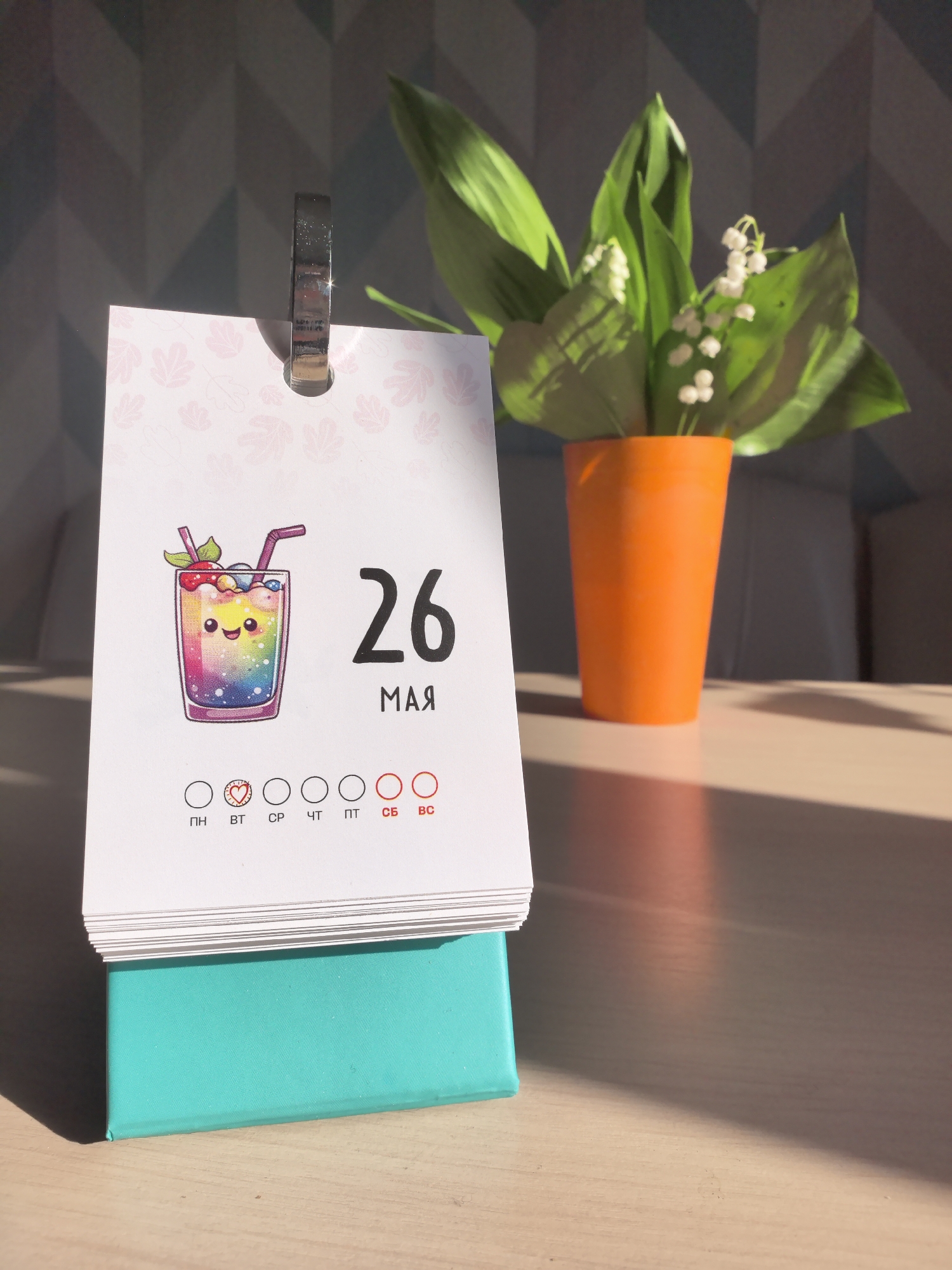 The image shows a calendar sitting on a light brown surface, likely a table, with a vase of flowers behind it. The calendar is open to the date May 26th. The calendar has a picture of a colorful cartoon drink, and the days of the week are written at the bottom. The base of the calendar is a light teal color. The flowers are lilies of the valley in an orange vase. The background is a wall with a subtle geometric pattern, partially obscured by sunlight. The location is the city of Фролы, Russia.