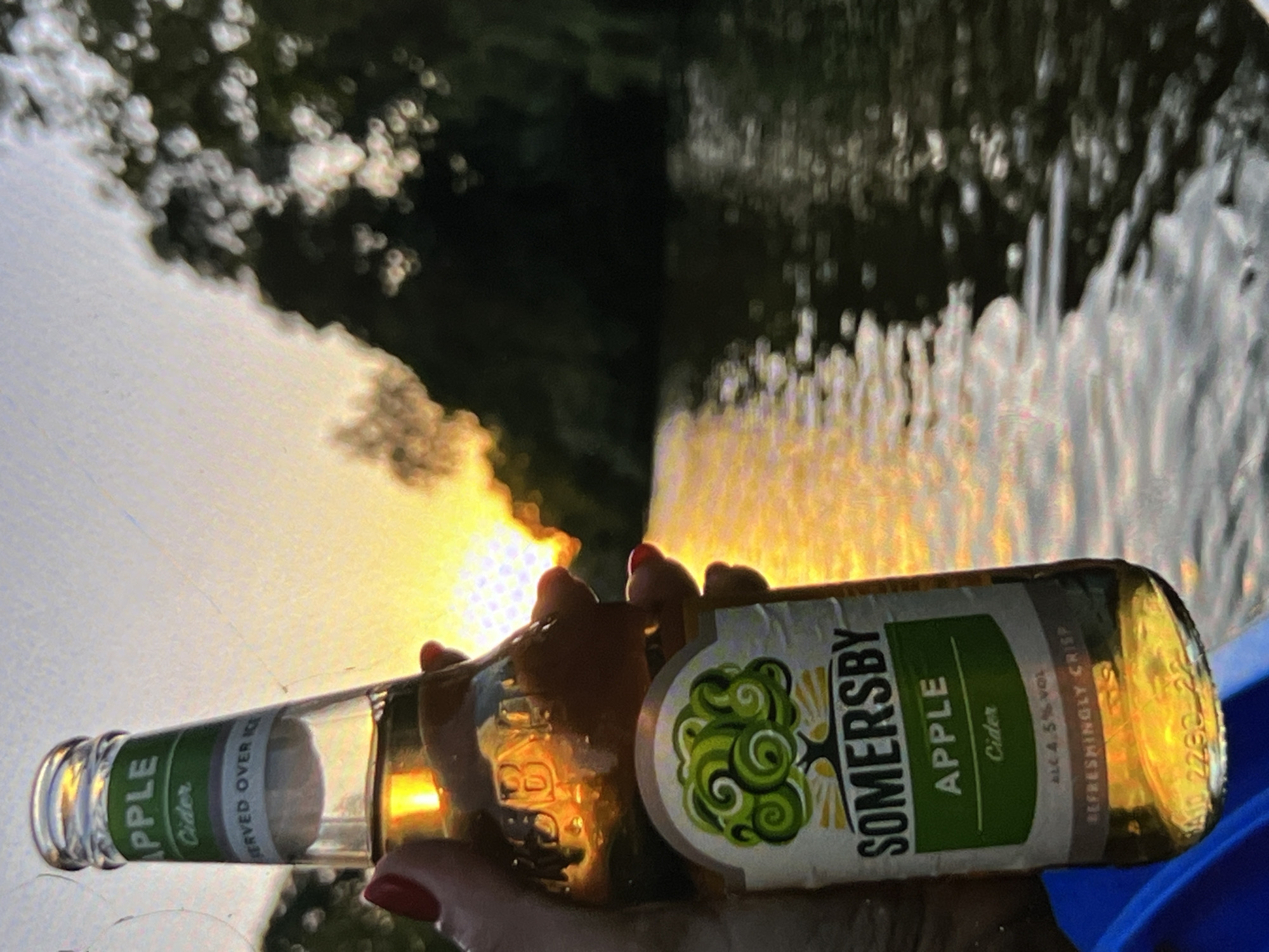 The image shows a hand holding a bottle of Somersby Apple Cider. The bottle is positioned horizontally, with the label facing the viewer. The label displays the brand name "SOMERSBY" and the flavor "APPLE Cider." Text indicating "ALC 4.5% VOL" and "REFRESHINGLY CRISP" is also visible on the label. The hand holding the bottle has red nail polish. In the background, the sun is setting, creating a warm glow, with blurred trees and water completing the backdrop. The city and state are Unknown.